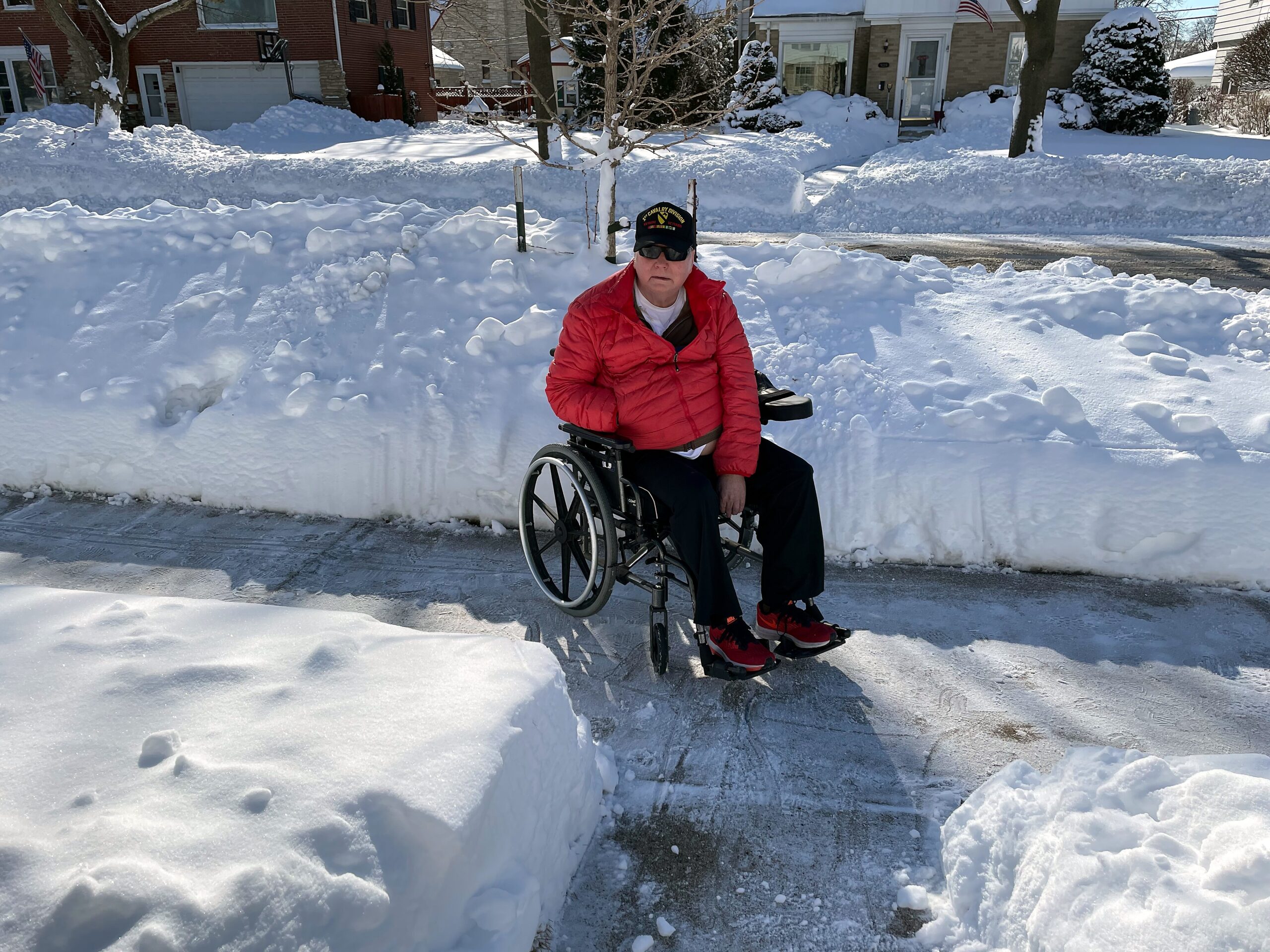 Person in a wheelchair in a red jacket on a snow-covered sidewalk with large snowbanks and houses in the background.