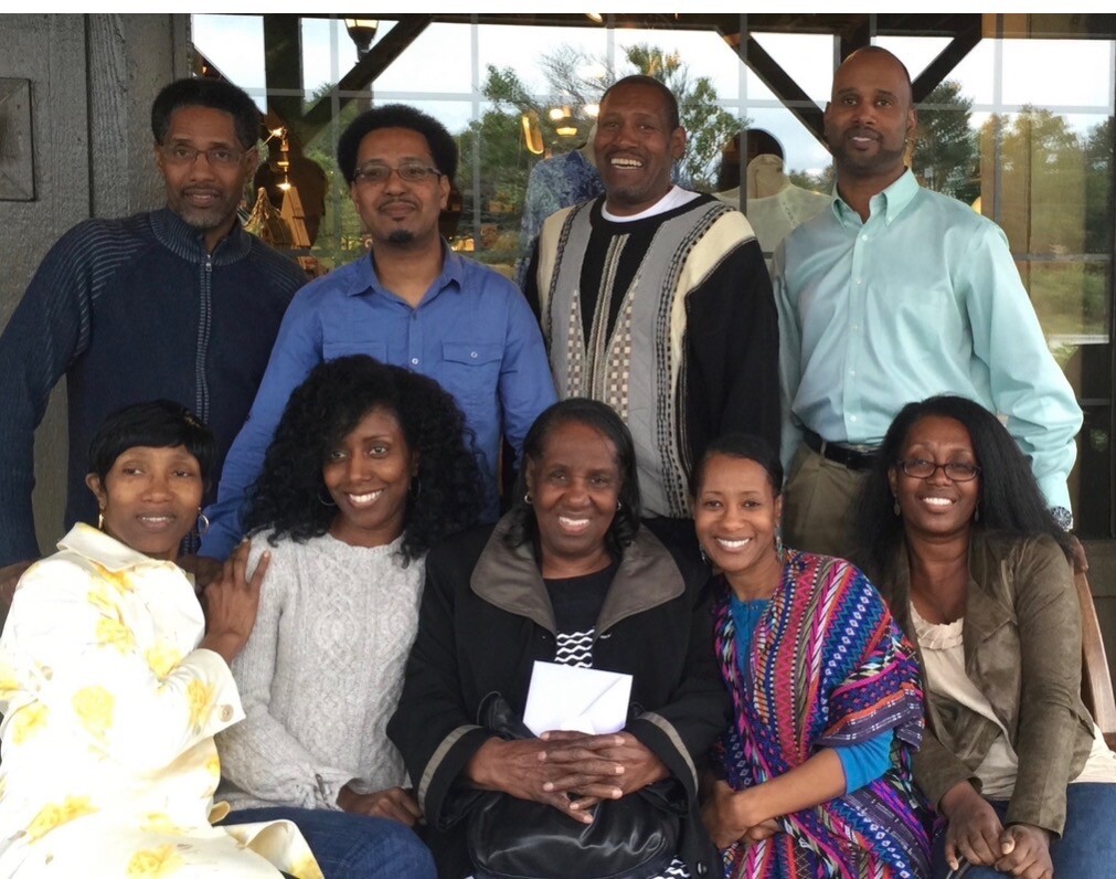 Nine adults posing for a group photo, five standing in back and four seated, smiling at the camera.