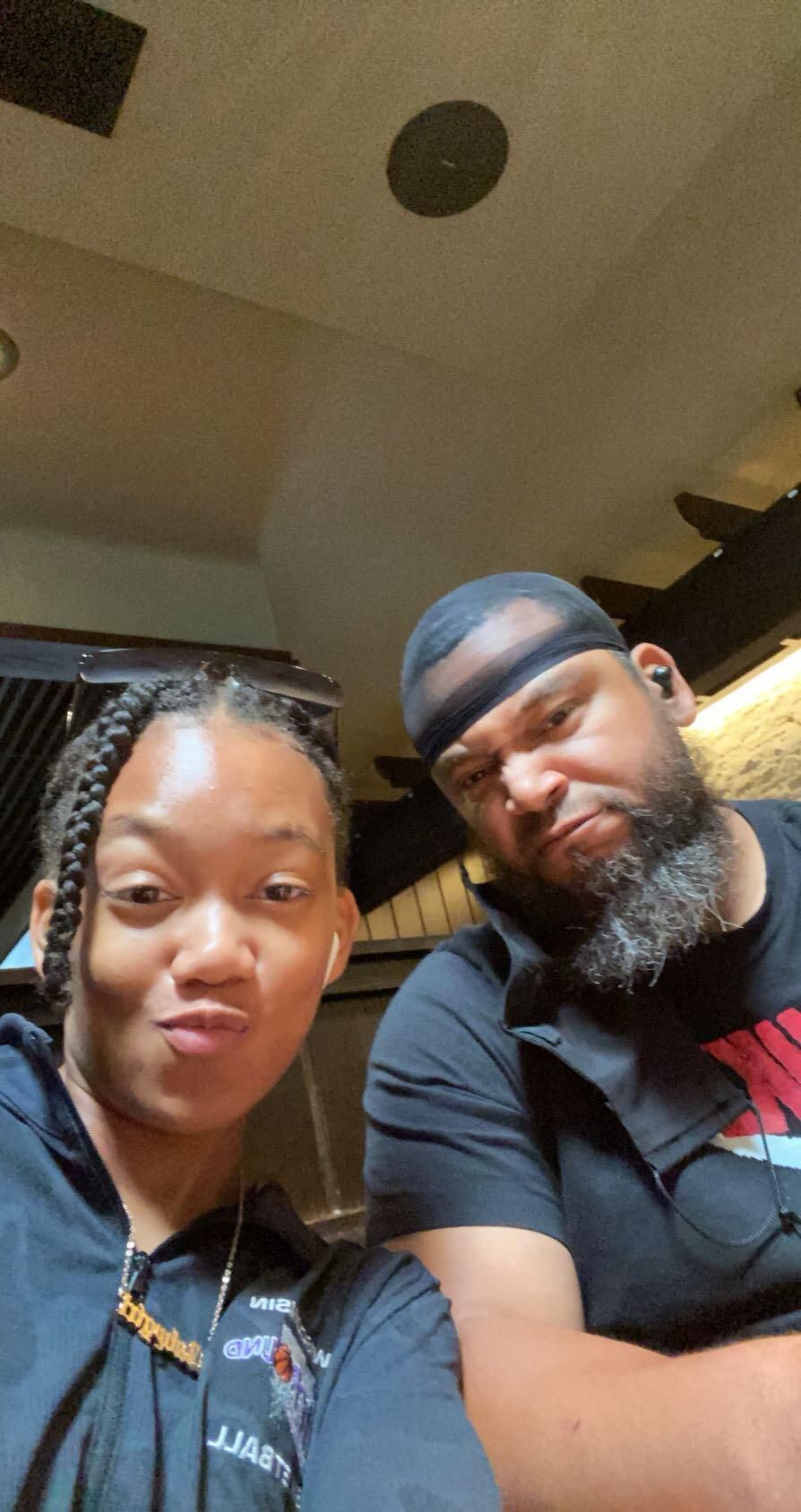 Two friends pose for a selfie in a cafe; woman with braids and sunglasses on her head smiles at the camera, man with a durag and beard beside her.