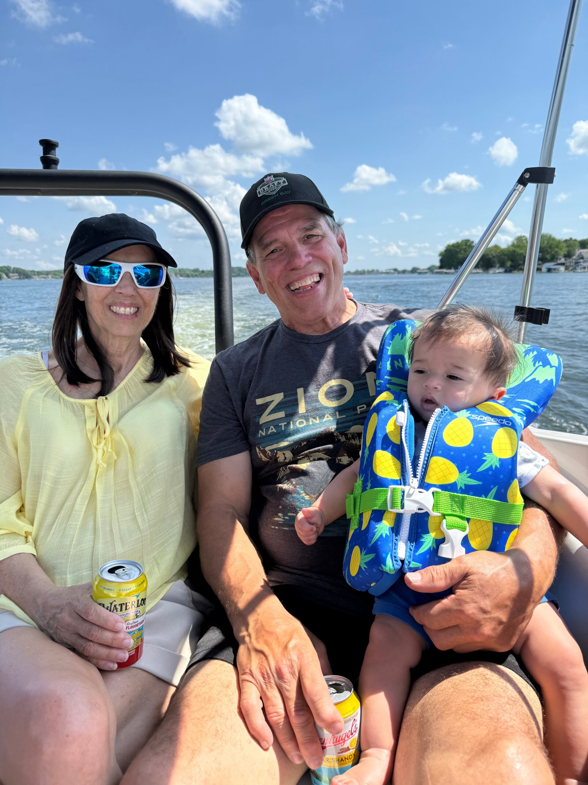 Smiling family on a boat: woman in a yellow blouse, man in a cap, and a toddler in a bright pineapple-pattern life jacket against a sunny lake backdrop.