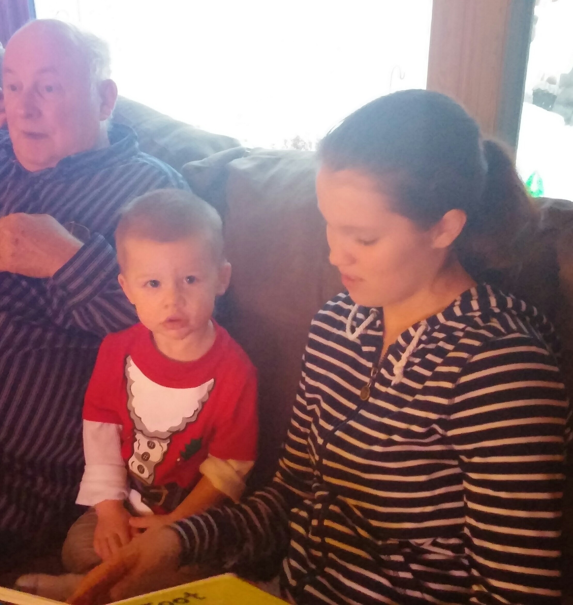 Older man with gray hair sitting on a dark couch next to a young boy in a red Santa shirt, with another child in a striped shirt nearby indoors.
