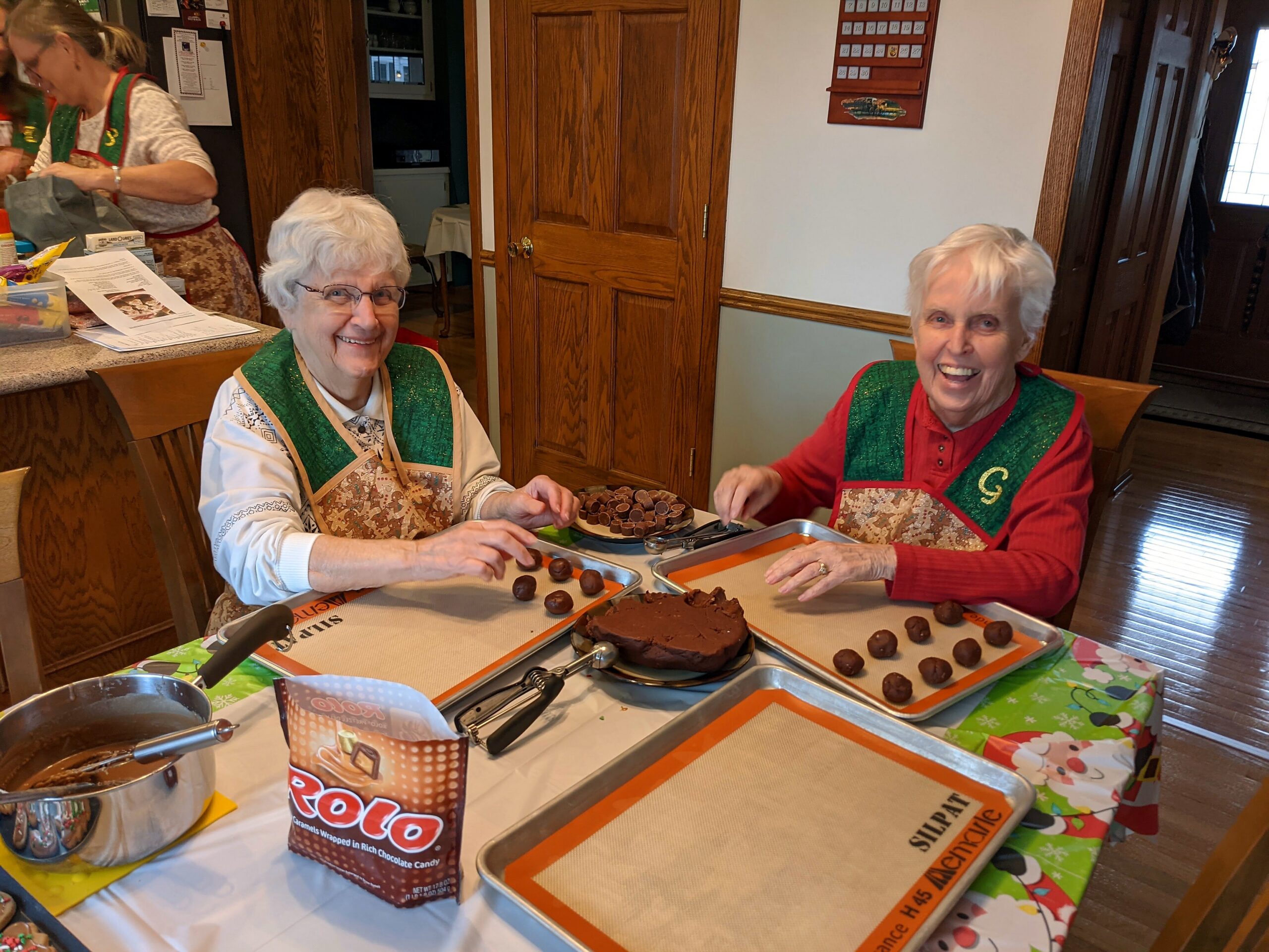 Two smiling elderly women in festive green aprons prepare chocolate candies at a holiday bake sale table.