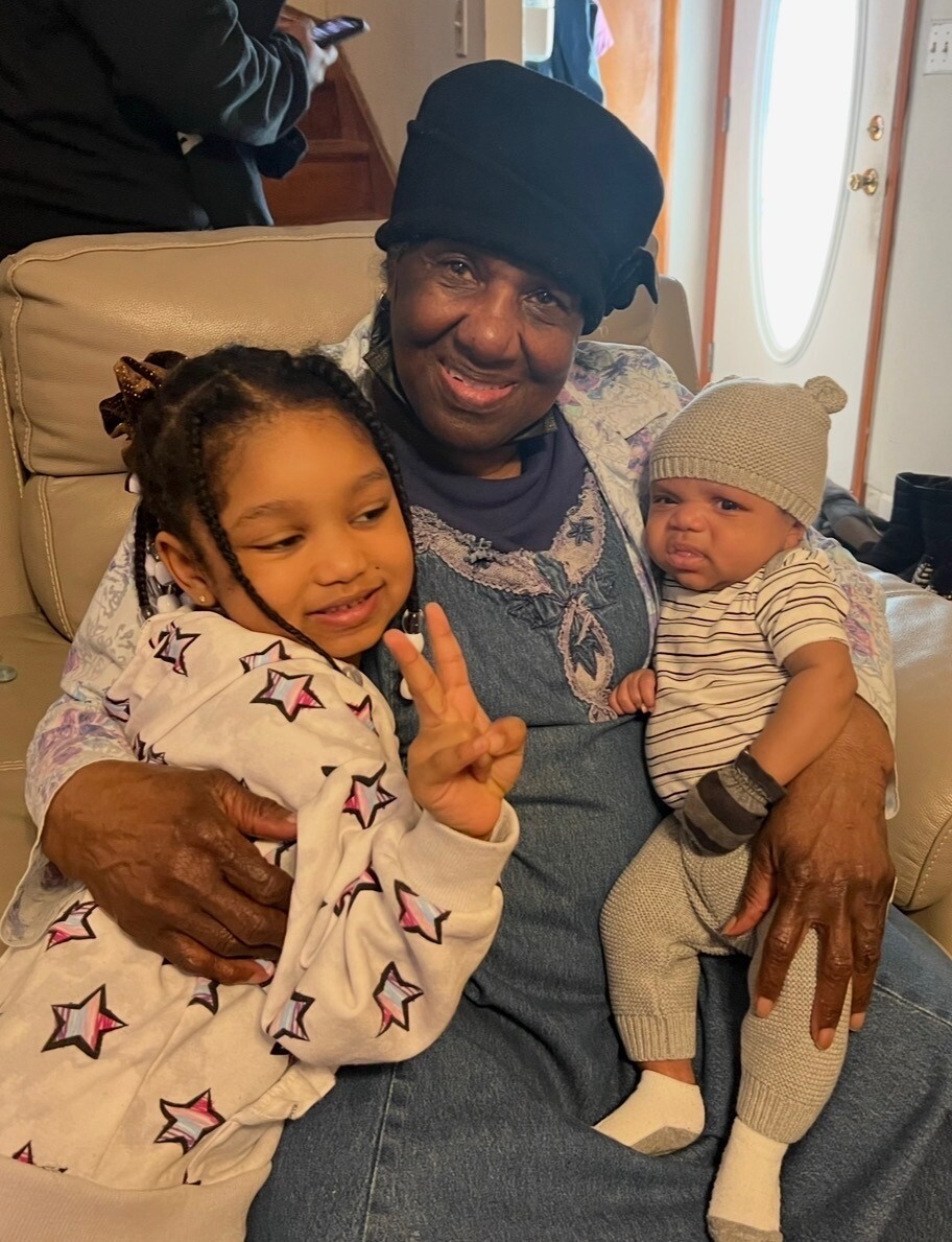 Older woman wearing a black hat sits on a beige sofa with a smiling baby and a young girl flashing a peace sign in a living room setting.