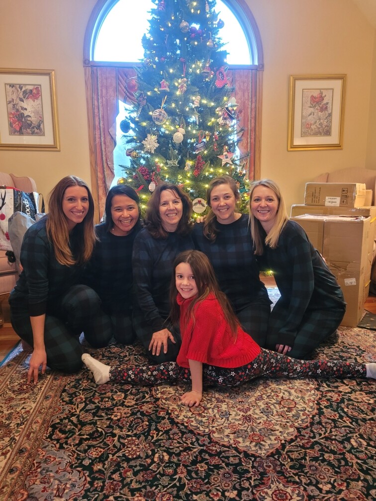 Six people pose for a Christmas photo in a living room: five women in matching plaid outfits and a girl in a red sweater doing a split on a patterned rug in front of a decorated Christmas tree with lights and ornaments and presents nearby.