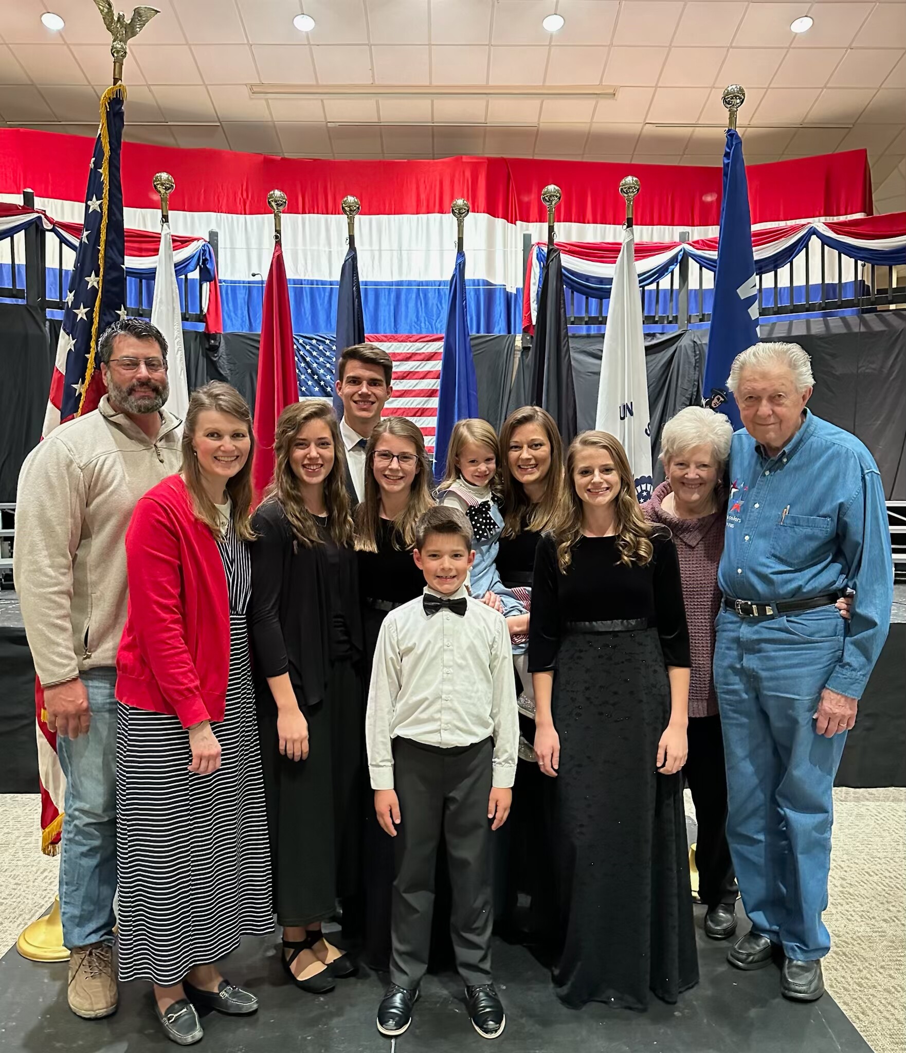 Smiling group of people of various ages posing on a stage with red, white, and blue banners and flagpoles behind them, including an American flag.