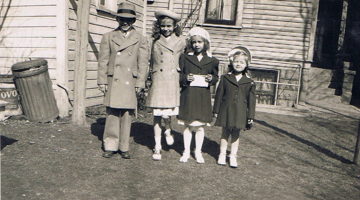 Four children stand in a dirt yard in front of a wooden house; two girls hold a white envelope or card.