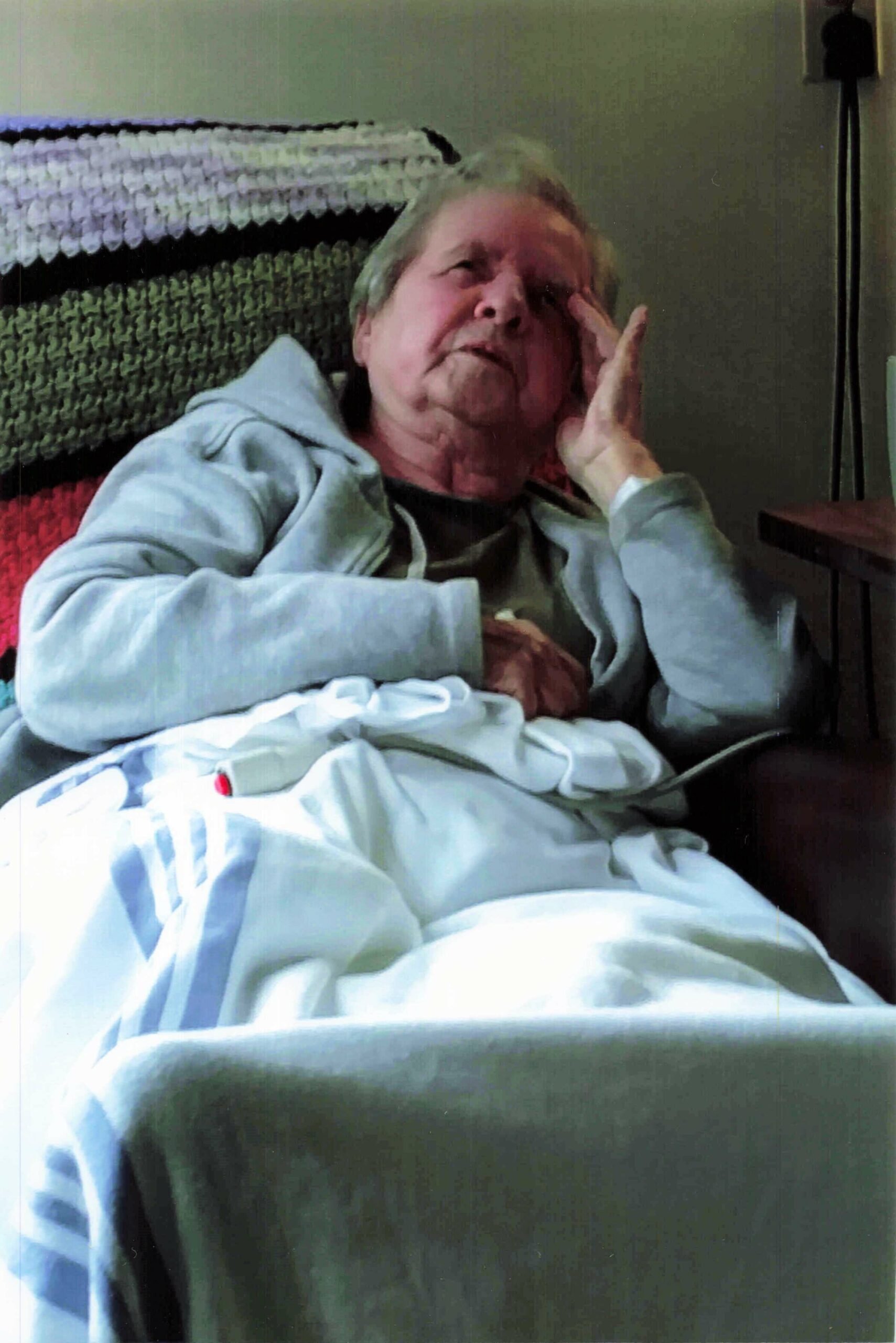 Elderly person resting in bed, hand to their cheek, wearing a light blue hospital gown with blankets around them in a dim room.