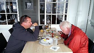 Two men sit at a light wooden table in a cafe, eating from bowls with drinks and condiments on the table nearby, engaging in conversation.