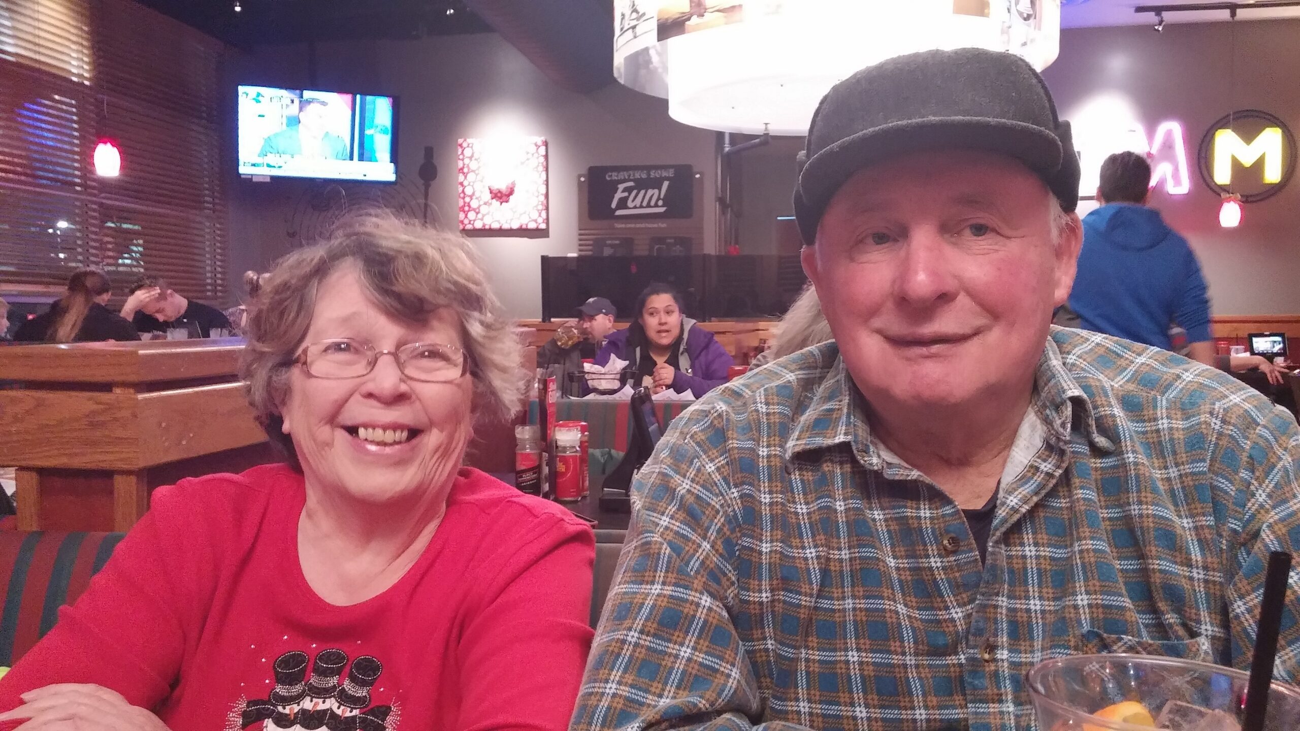 Smiling older couple seated at a restaurant table; woman in a red top on the left, man in a plaid shirt and cap on the right, with other diners and neon signage in the background.