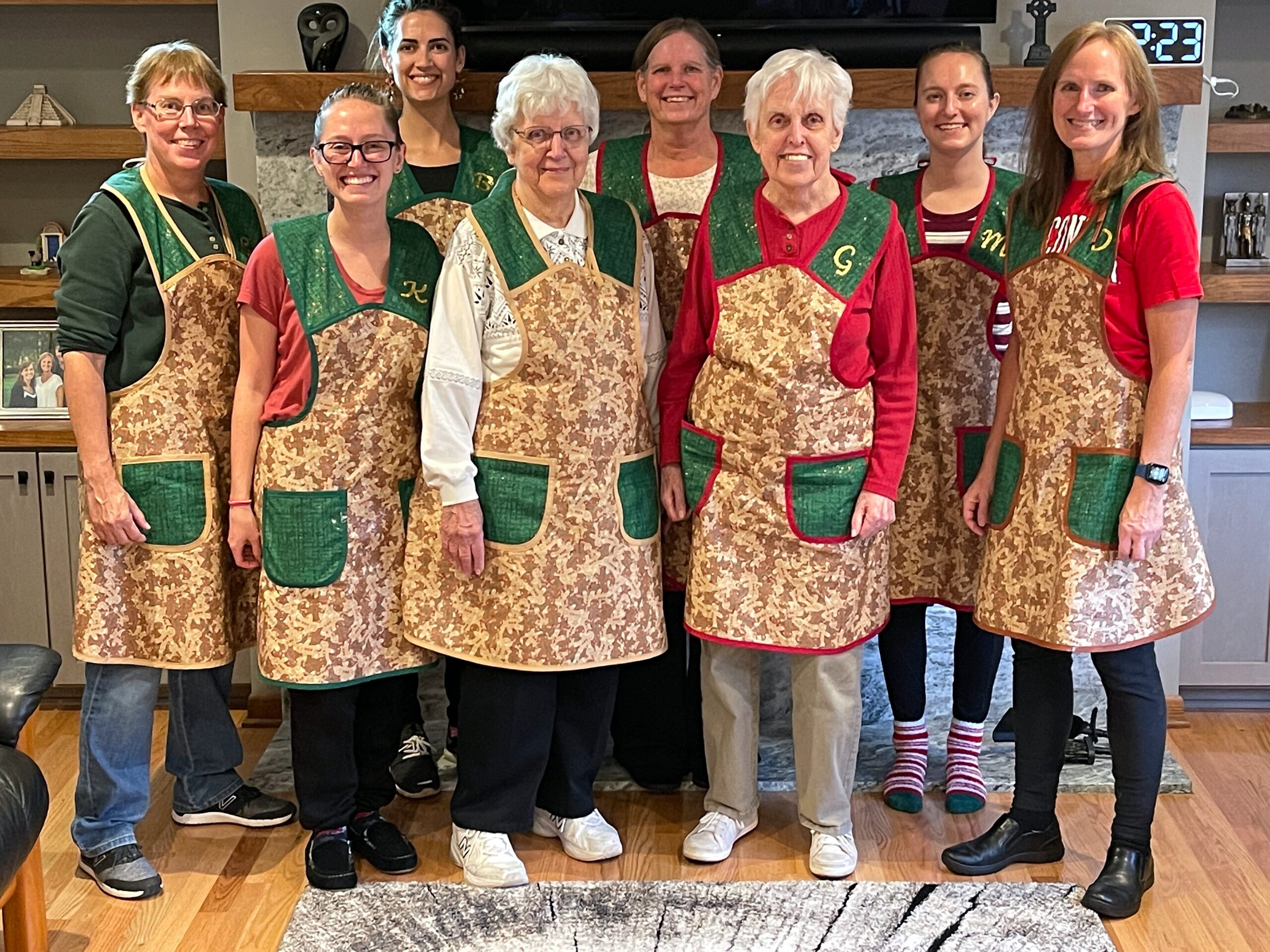 Group of women wearing matching brown floral aprons standing together in a kitchen, smiling at the camera.