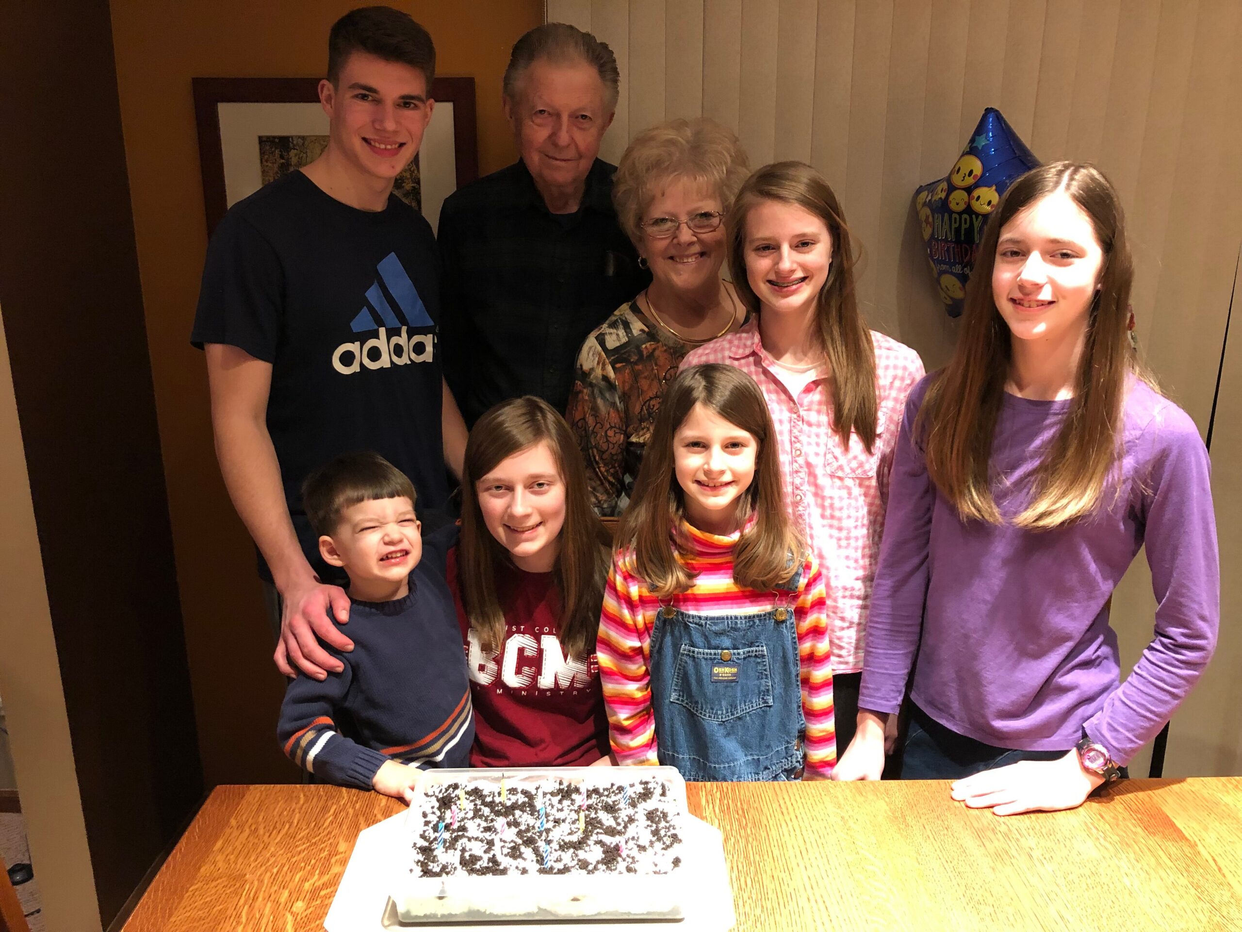 Smiling family gathered around a table with a decorated birthday cake and a birthday balloon in the background.