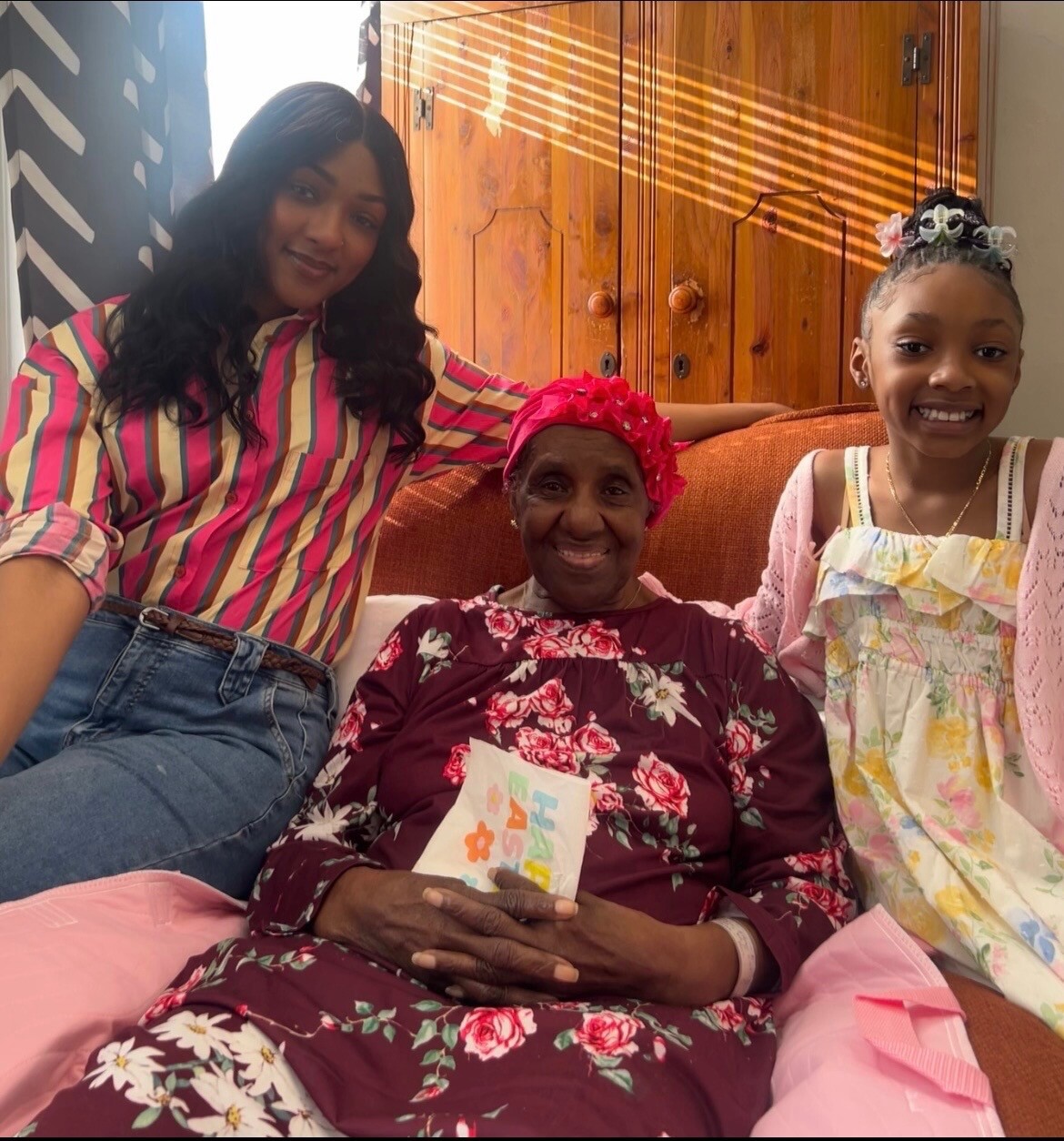 Three generations posing together on a couch: an elderly woman in a pink headwrap and floral dress, flanked by two younger women smiling warmly.