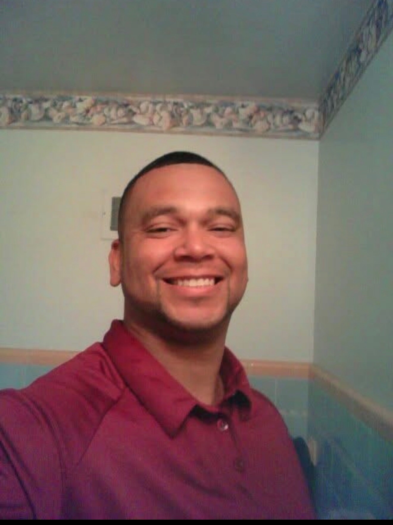 Smiling man in a maroon shirt taking a selfie in a room with pastel walls and wallpaper border to the ceiling