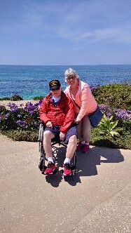 Two older adults pose together by the seaside on a sunny day; one person sits in a wheelchair wearing a red jacket and black cap, with a companion in a pink jacket beside them, purple flowers and blue water in the background.