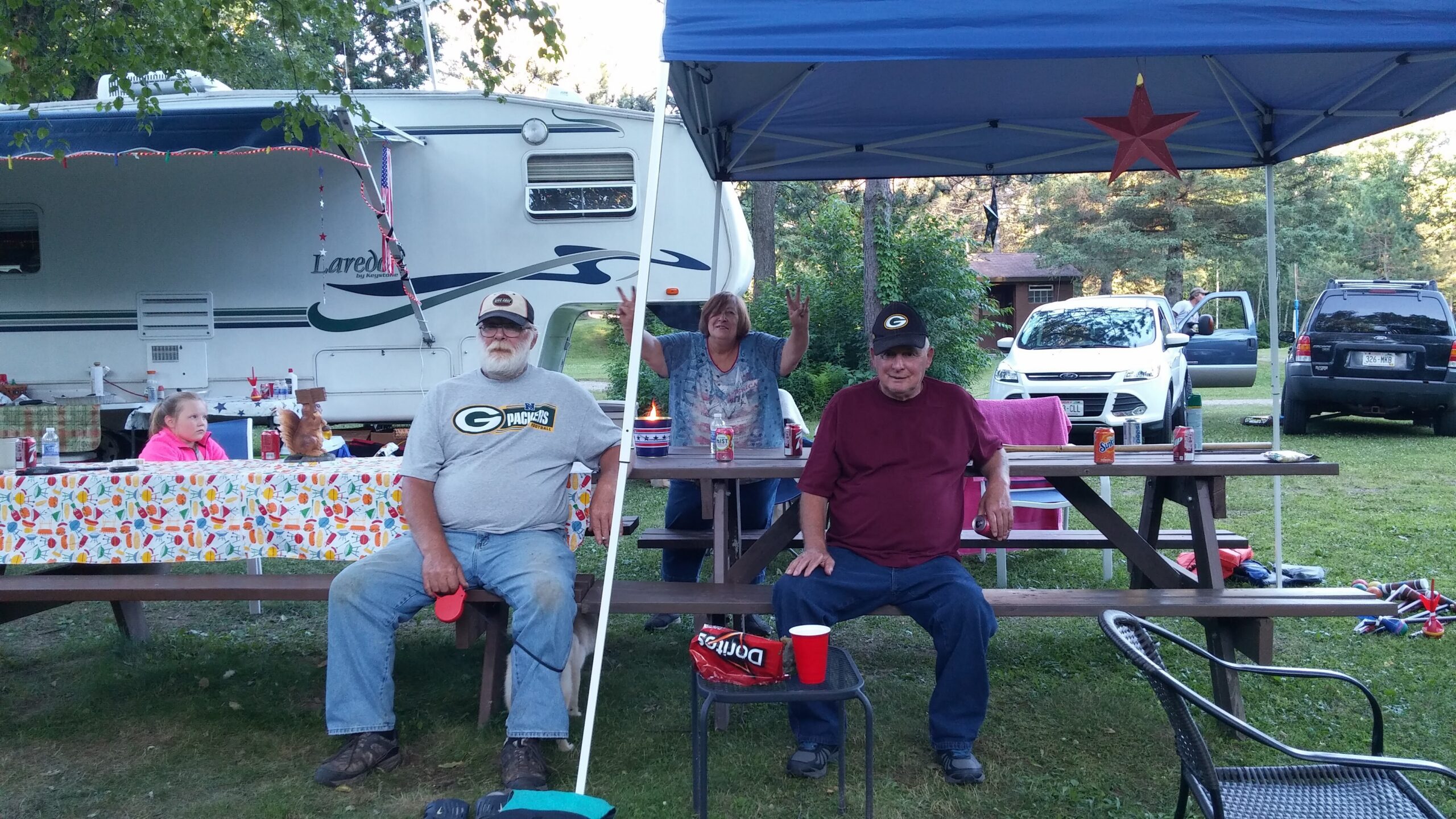 Family gathering at a campground with a large white RV, a blue canopy, and people seated at picnic tables outdoors under trees.