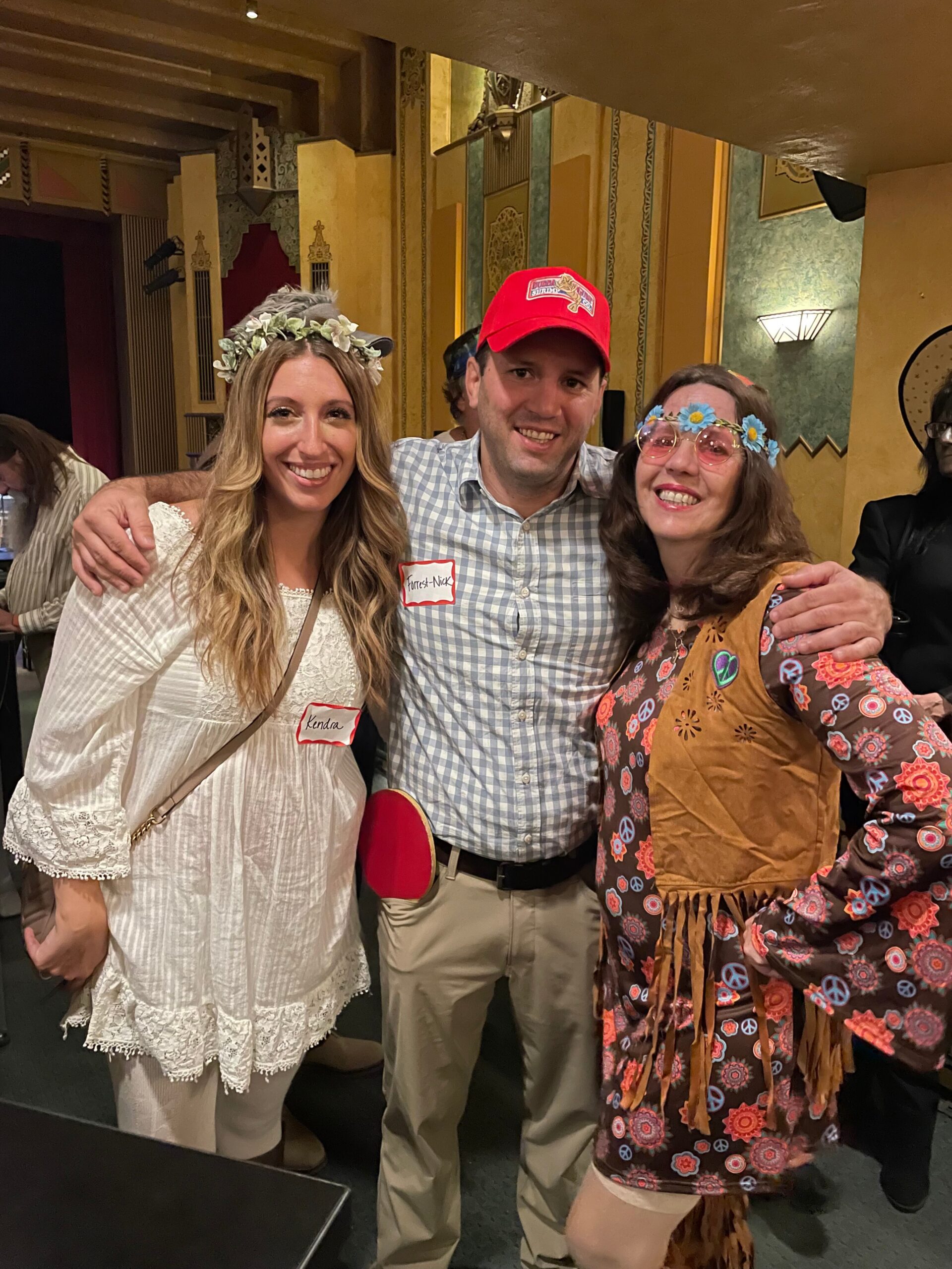 Three people in vintage costumes pose together indoors: a woman in white lace with a flower crown, a man in a check shirt and red cap, and a woman in a floral fringe dress with blue flower headband.