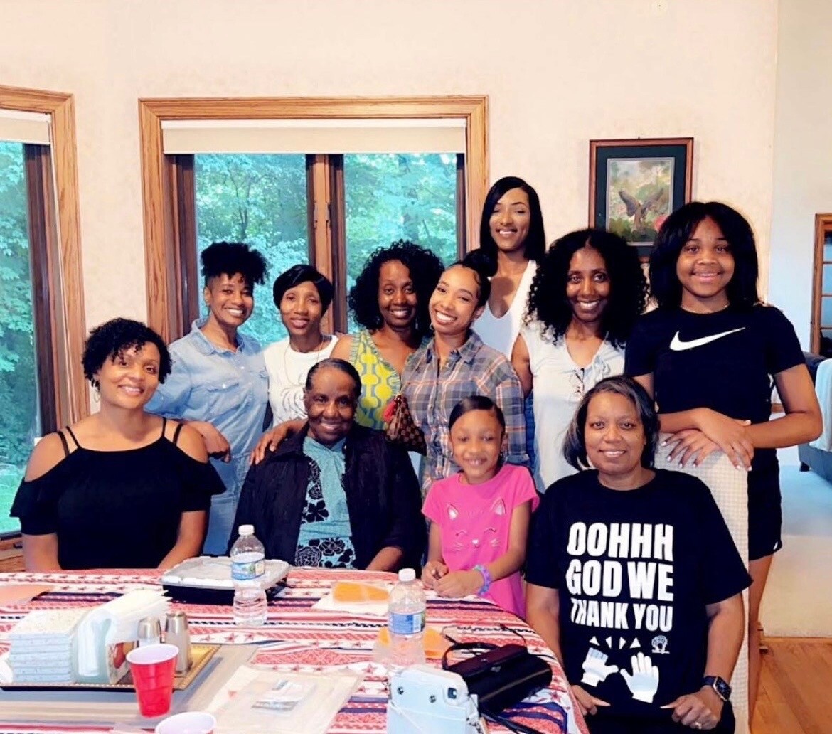 A diverse group of adults and a child pose together around a decorated table in a sunlit living room, smiling for the camera.