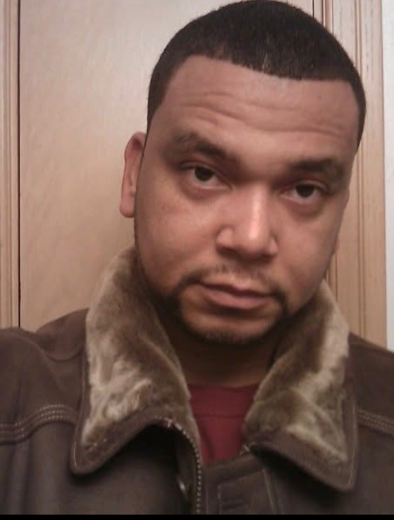 Close-up portrait of a man with short dark hair and light facial stubble, wearing a brown jacket with a furry collar and looking at the camera.