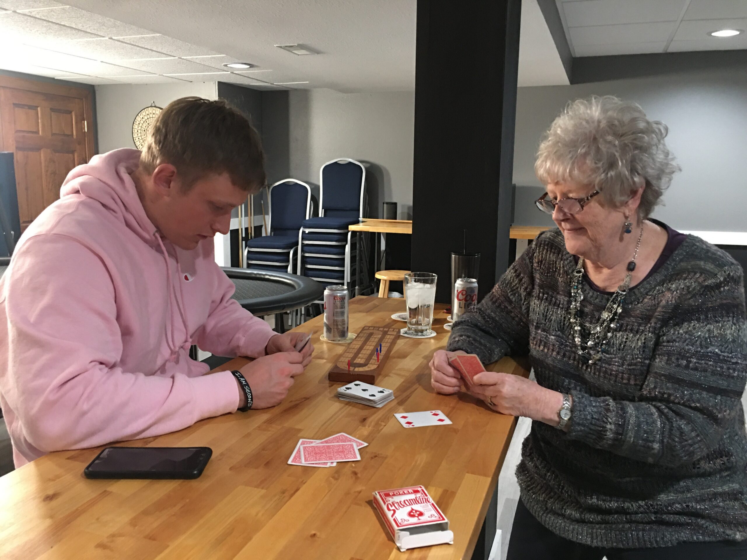 Two people sit at a wooden table playing cards, focused on their hands. A younger person in a pink hoodie and an older woman with glasses share the game in a basement setting.