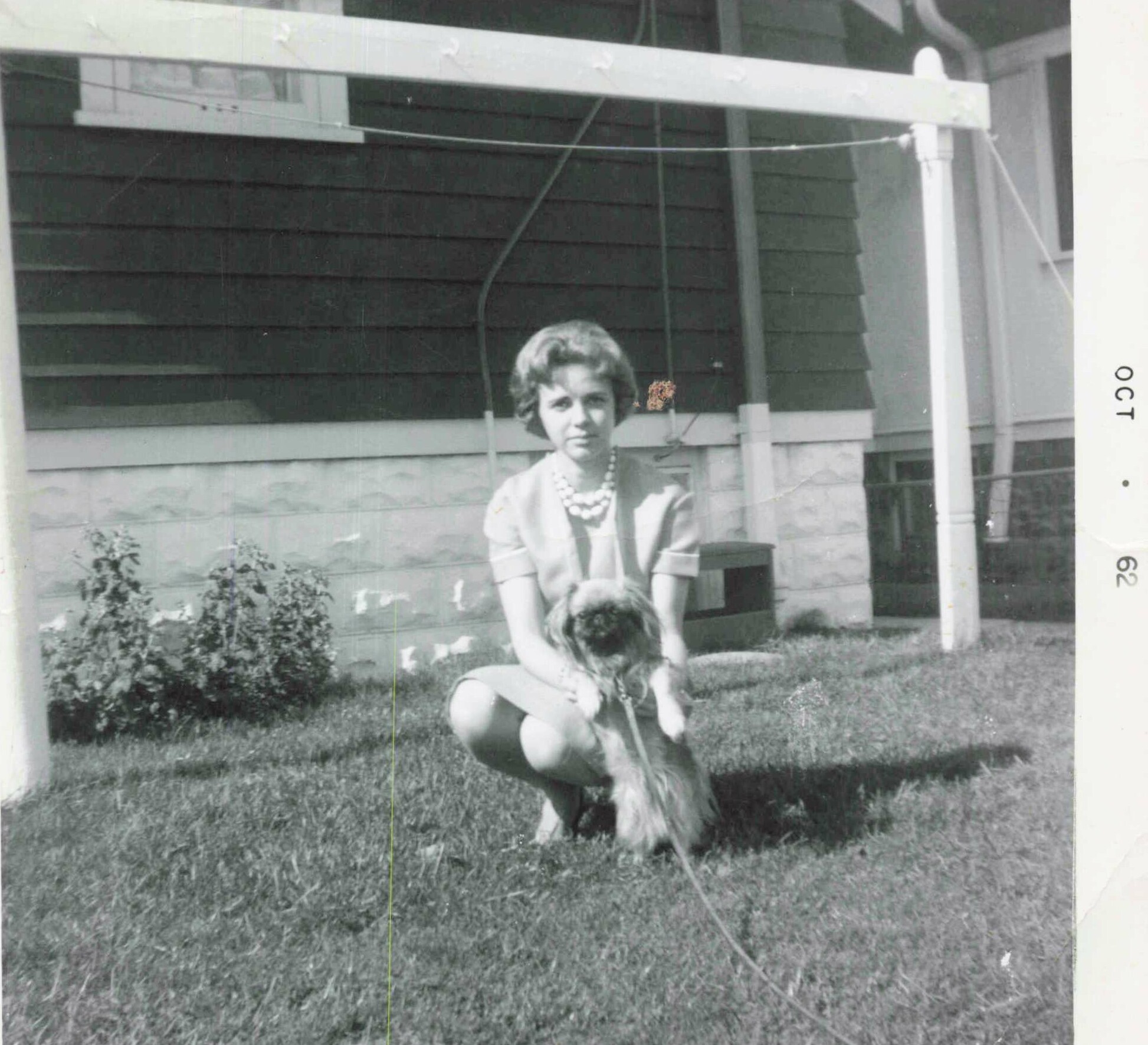 Woman in a light dress and pearl necklace kneels in a front yard beside a small fluffy dog in front of a house porch.