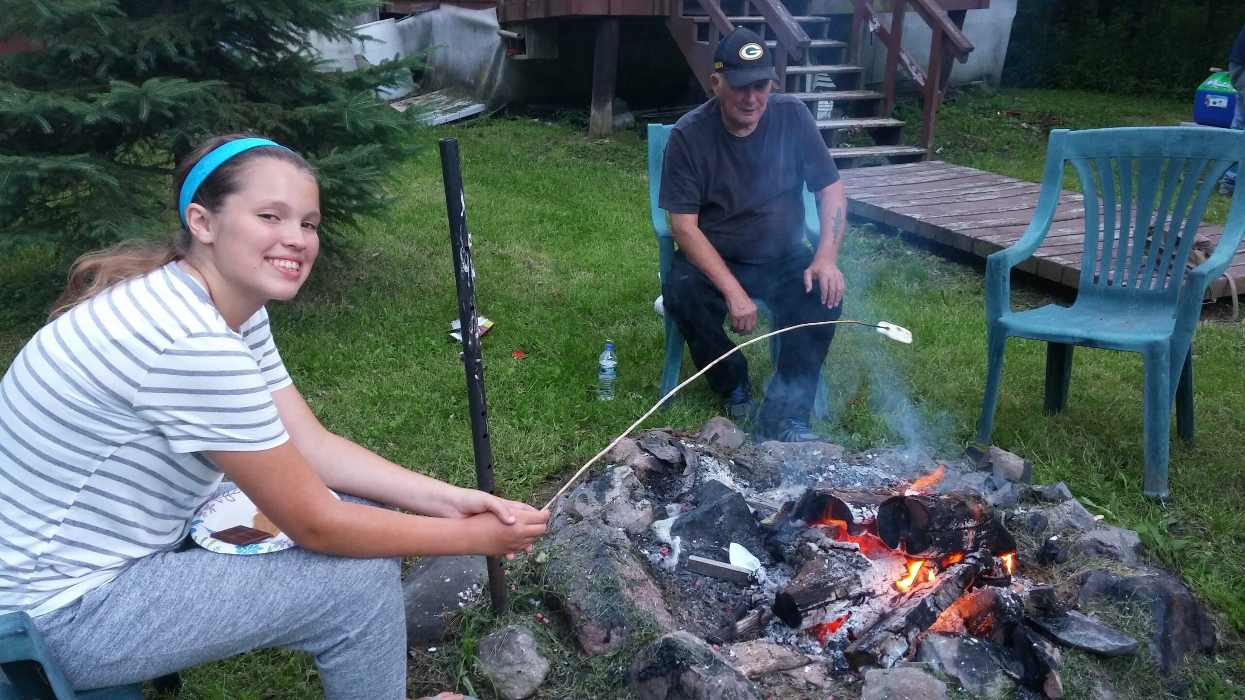 Girl with a blue headband and striped shirt smiles while roasting a marshmallow over a backyard fire with a man nearby, seated on plastic chairs.