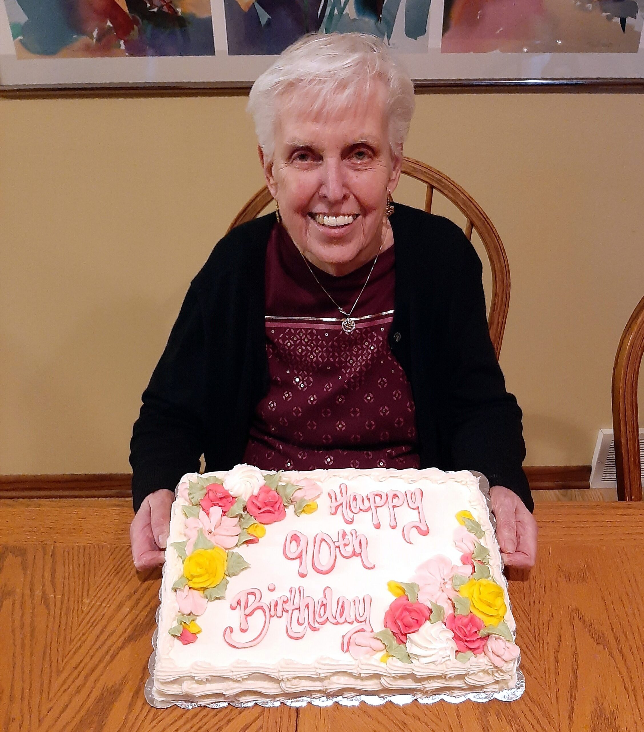 Smiling elderly woman with short white hair seated at a table with a large decorated birthday cake in front of her.
