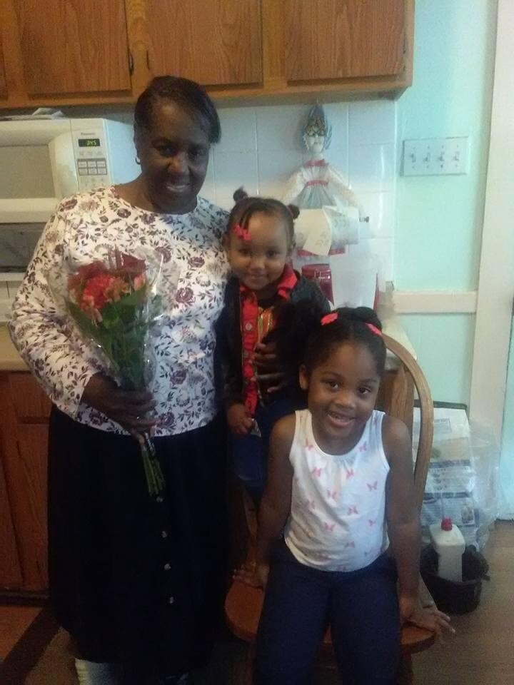 Smiling woman in a floral white top holding a bouquet of red flowers, accompanied by two children in a kitchen—girl in a white sleeveless top with pink butterflies sitting on a chair and boy in a red shirt standing behind her.