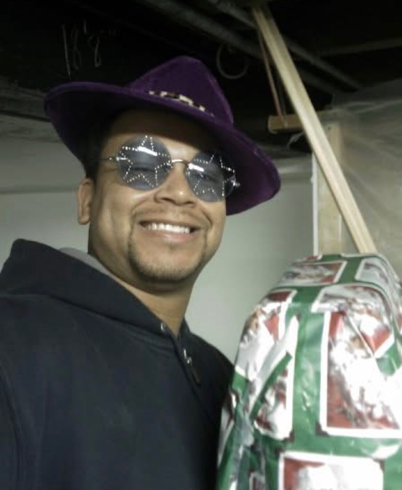 Smiling man in a purple hat and round sunglasses holding a wrapped gift in festive green and red paper.