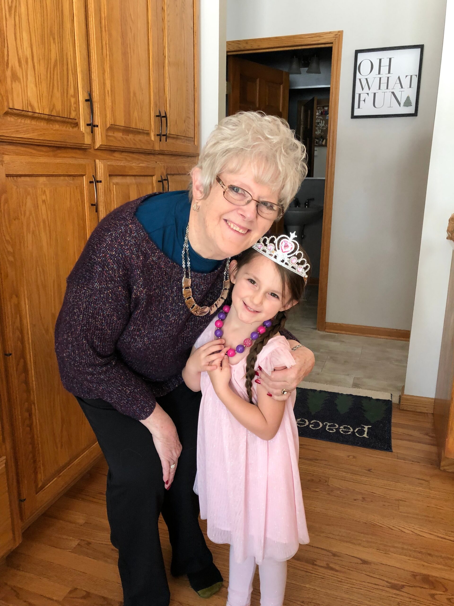 Smiling elderly woman in a dark purple sweater hugs a young girl in a pink princess dress and tiara in a home entryway.