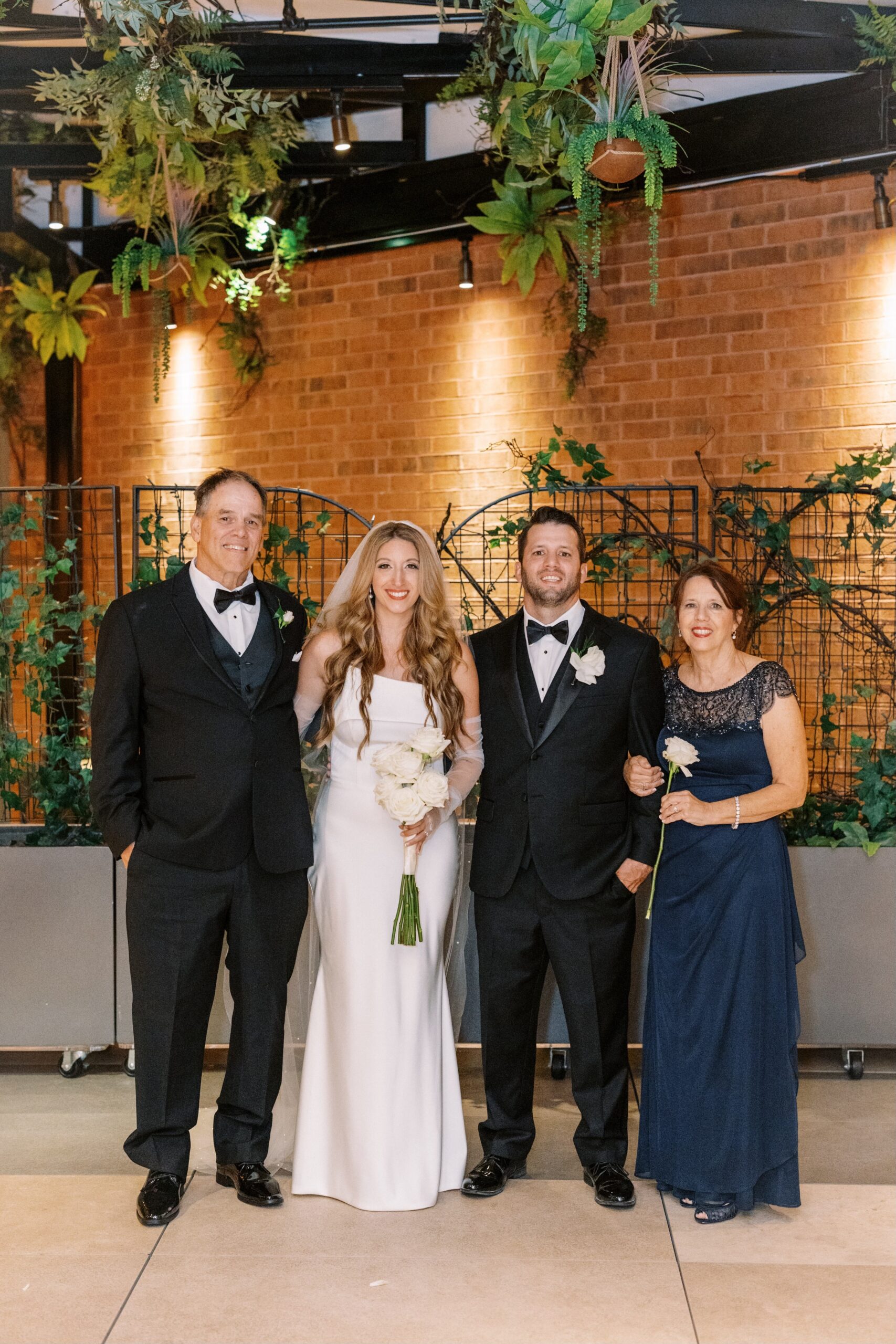 Bride in a white gown holding a bouquet stands with two men in black tuxedos and a woman in a navy blue dress indoors against a brick wall and greenery.