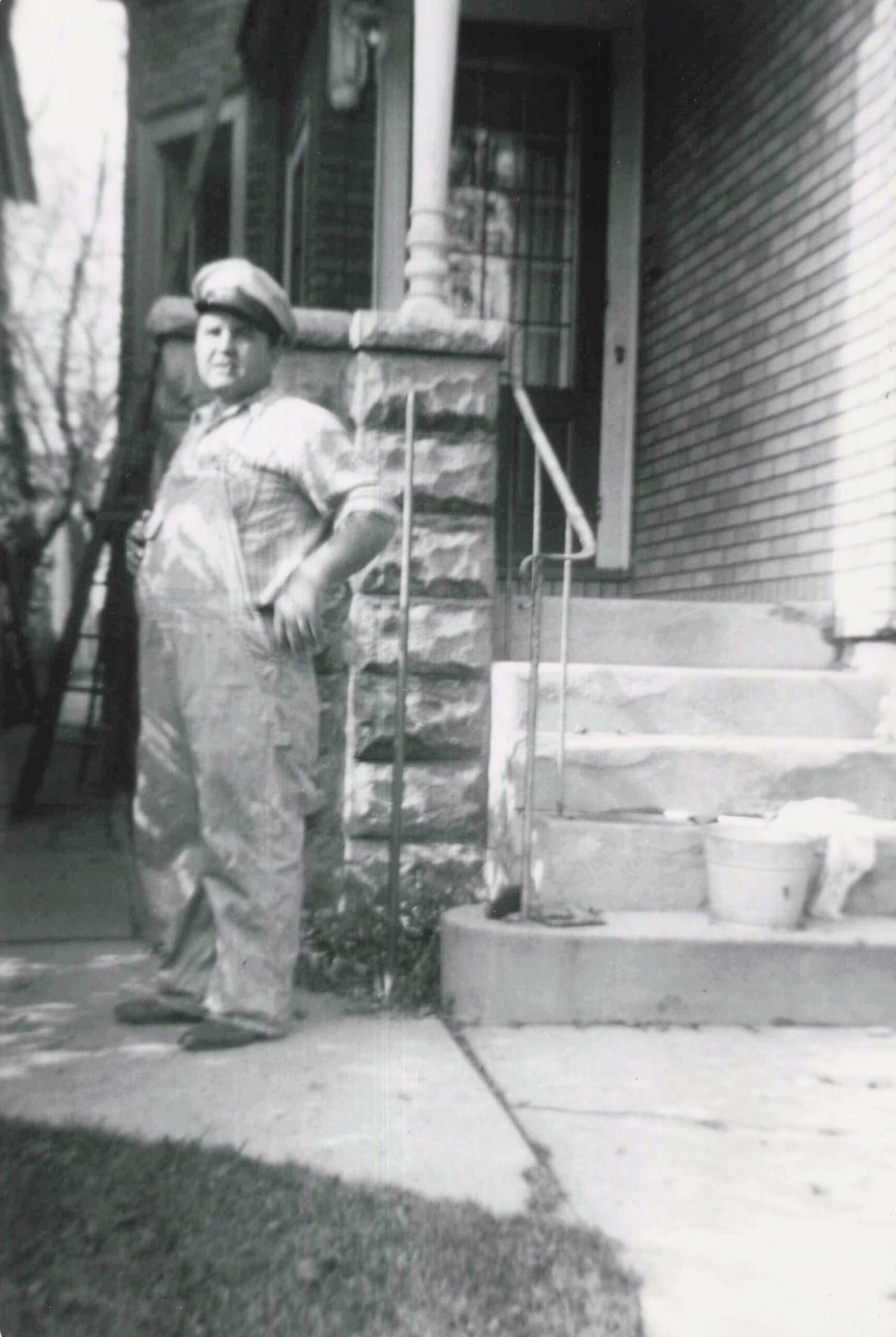 Man wearing a cap and work clothes standing on stone steps outside a brick house, near a railing and doorway (black-and-white photo).