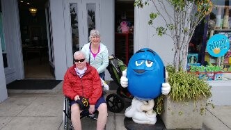 Two seniors pose outside a storefront with a blue cartoon mascot giving thumbs up; one man sits in a wheelchair wearing a red jacket, and a woman stands beside him smiling.