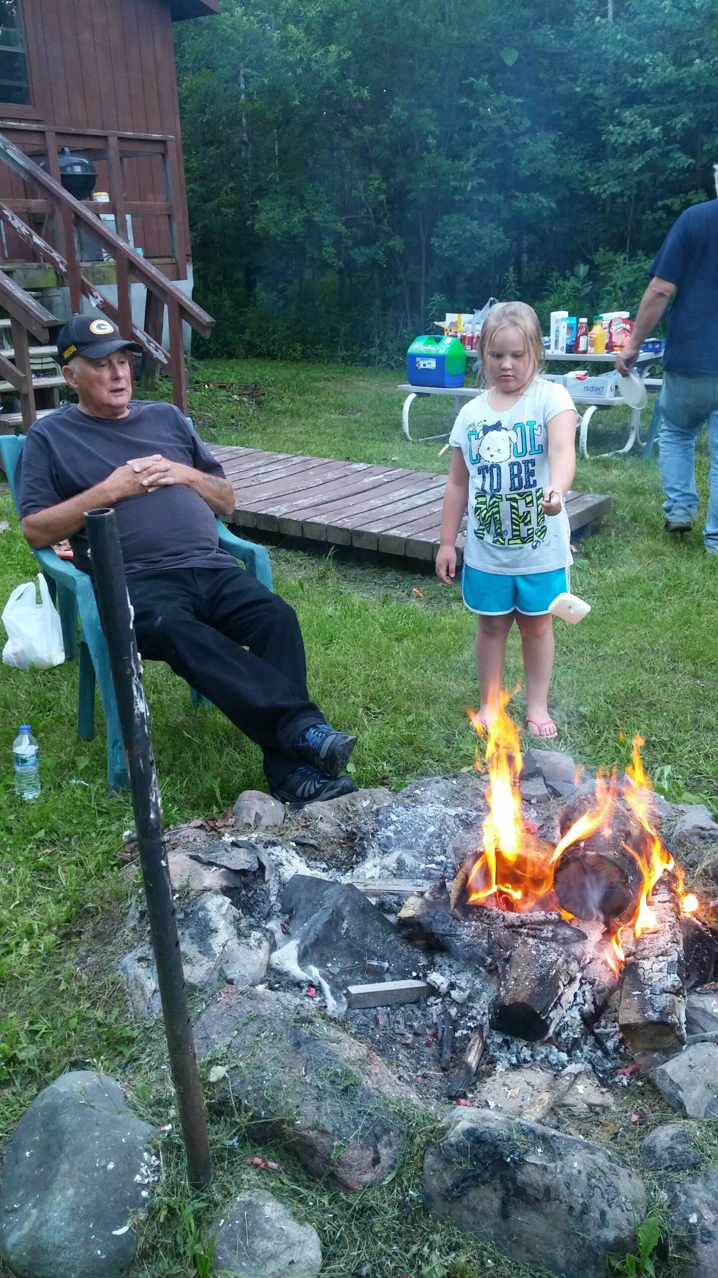 An elderly man wearing a cap sits in a green plastic chair beside a campfire, while a young girl in a white T-shirt and blue shorts stands nearby in a grassy backyard with a table of supplies in the background.