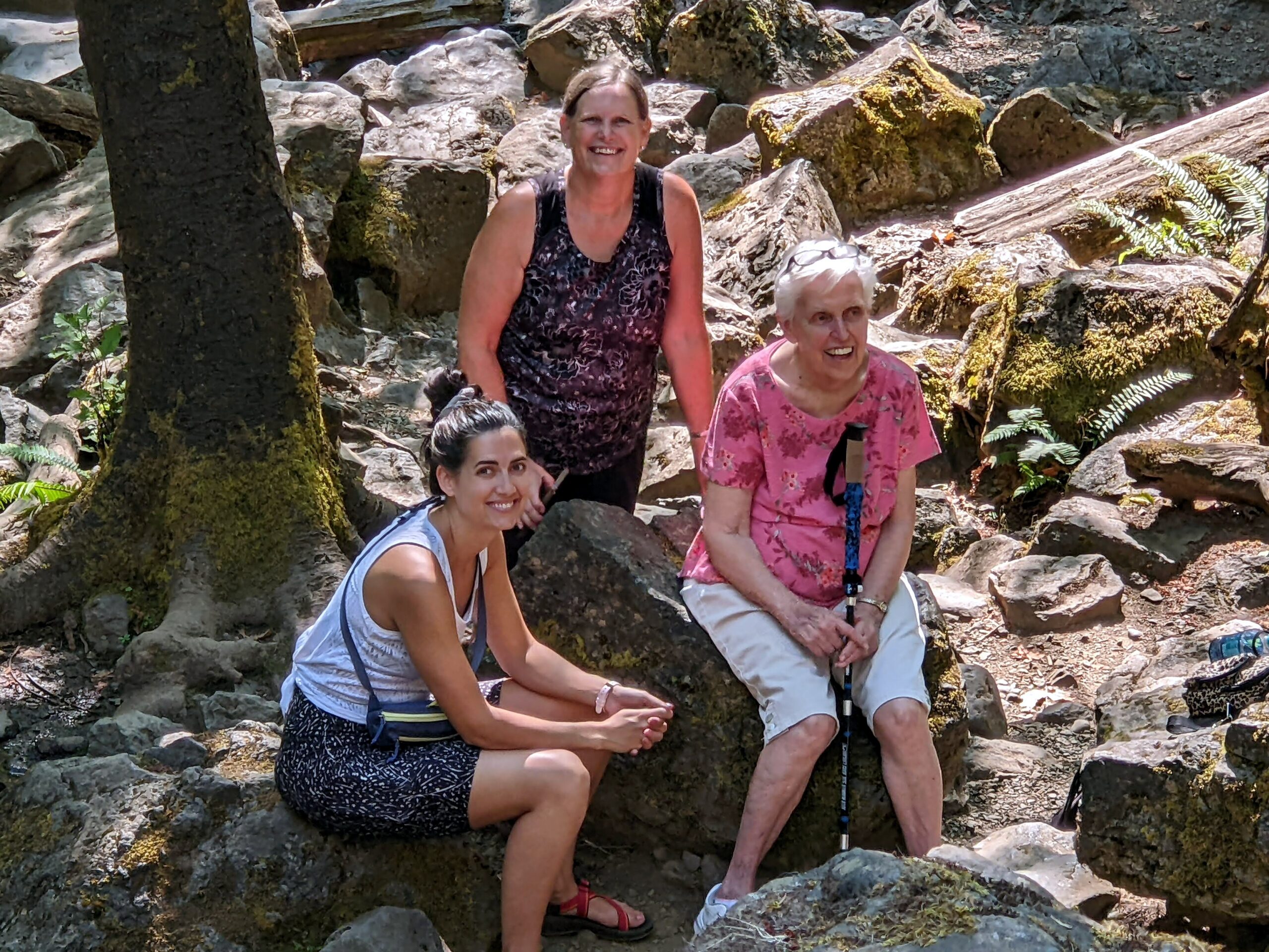 Three women pose on a rocky forest trail during a hike, wearing casual outdoor clothing and backpacks amid mossy rocks and ferns.