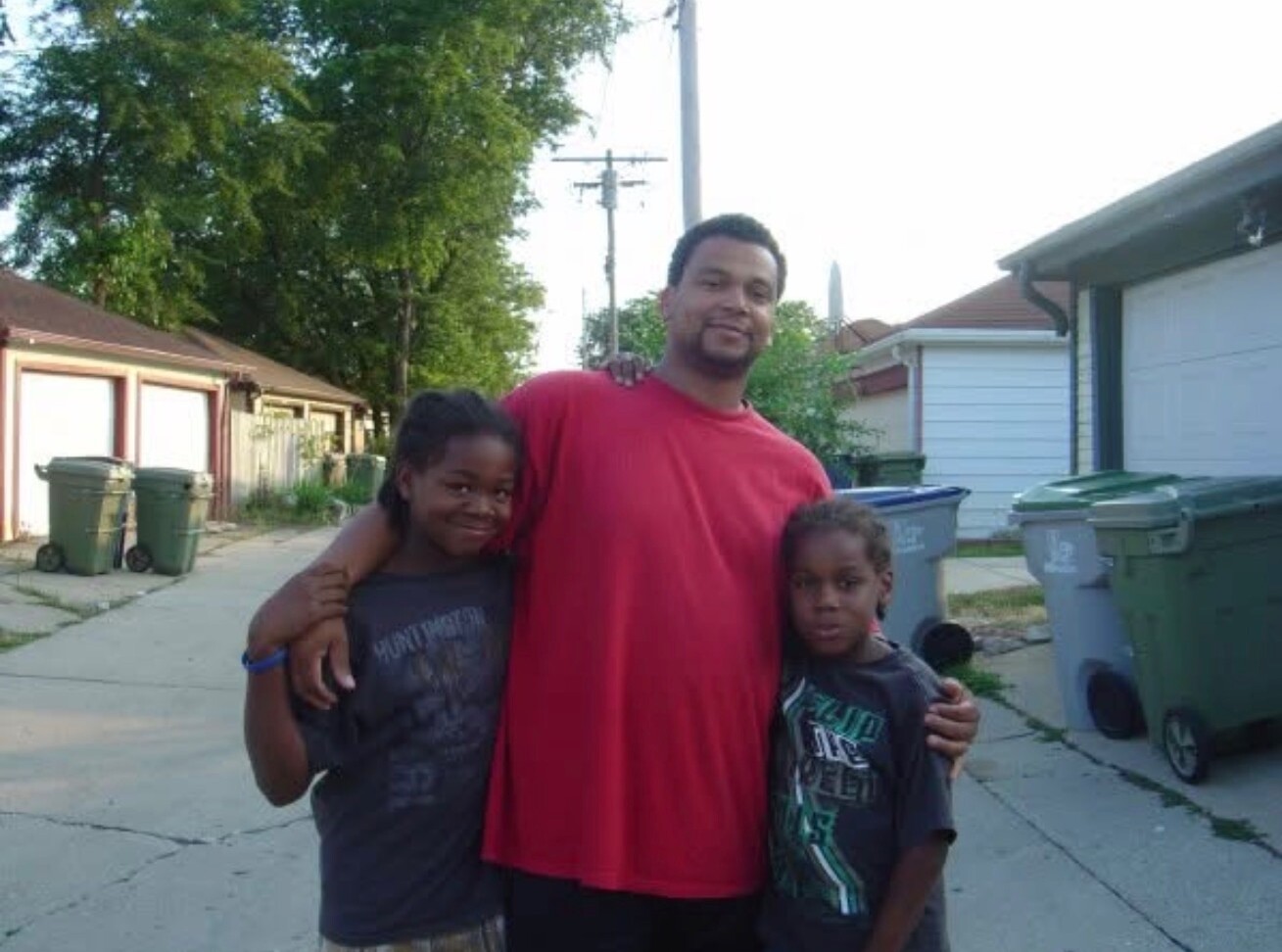 Man in red shirt with two children, arms around them, smiling in a residential driveway outdoors.