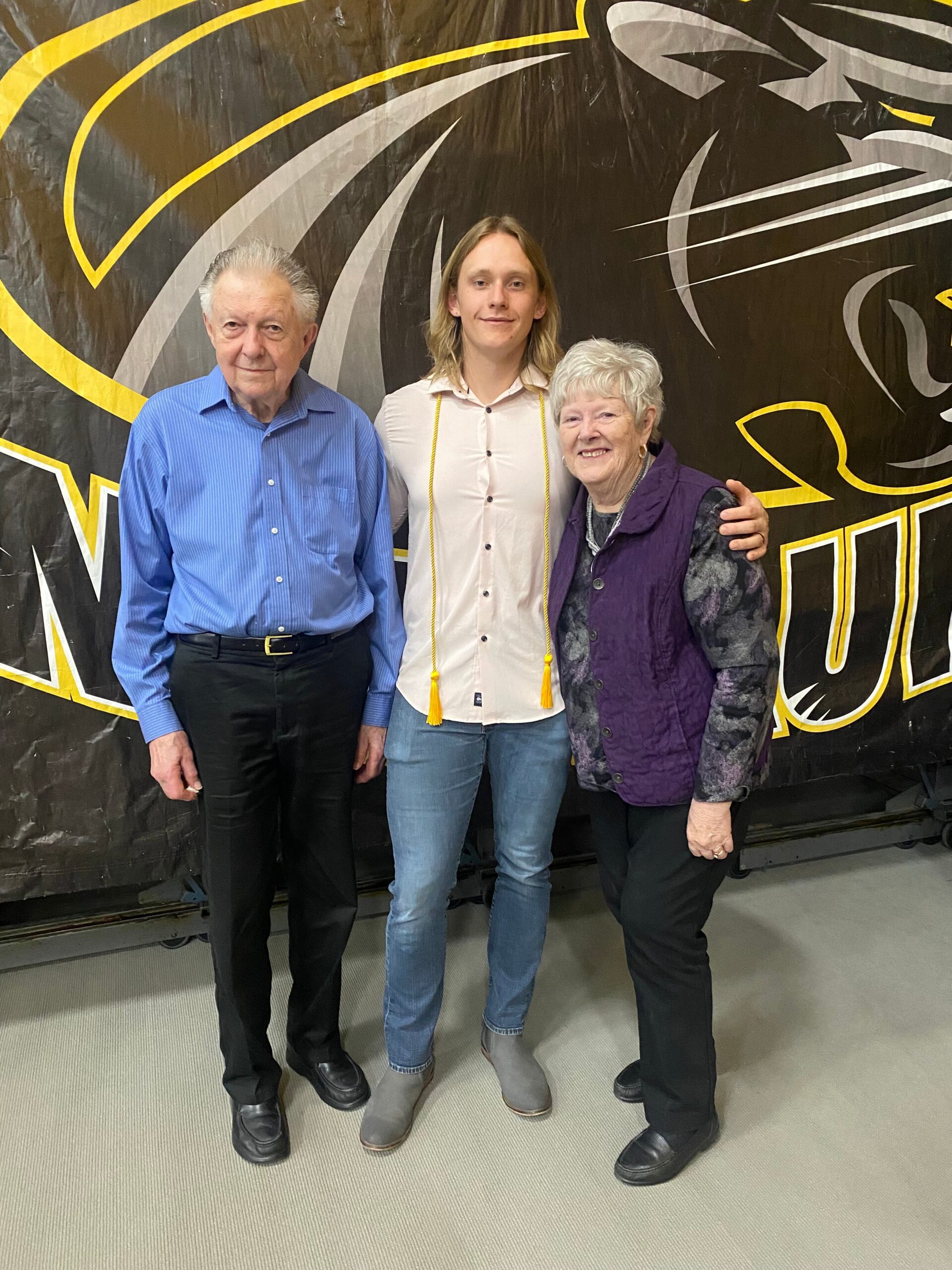 Three people pose for a photo in front of a black banner with yellow and gray graphics: an older man in a blue shirt, a young adult with graduation cords, and an older woman in a purple vest.