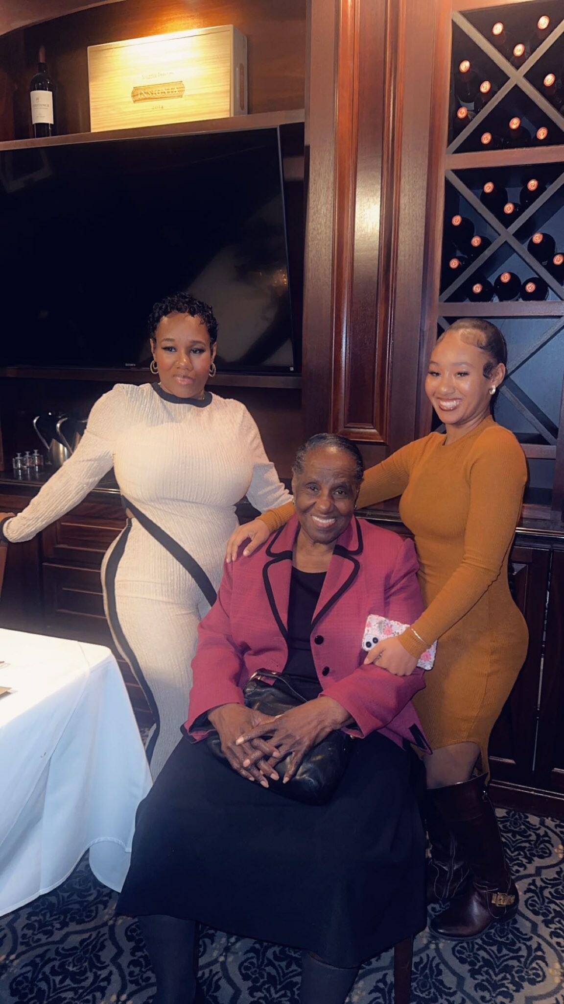 Three women posing together indoors: an elderly woman in a pink blazer seated between two smiling younger women, one in a white dress and the other in a mustard dress, with a wine rack and wooden cabinets in the background.