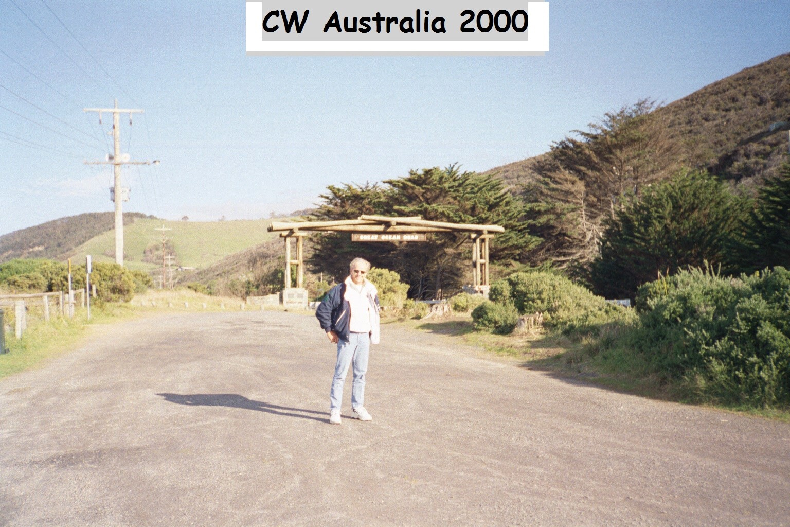 Man standing on a gravel road near a wooden entrance arch in a rural hillside setting.
