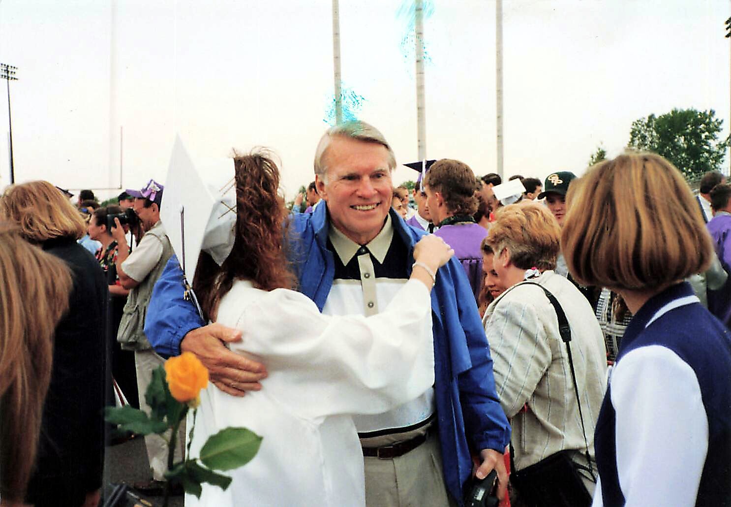 Graduate wearing a white gown hugs an older man in a blue jacket at a crowded outdoor graduation event.