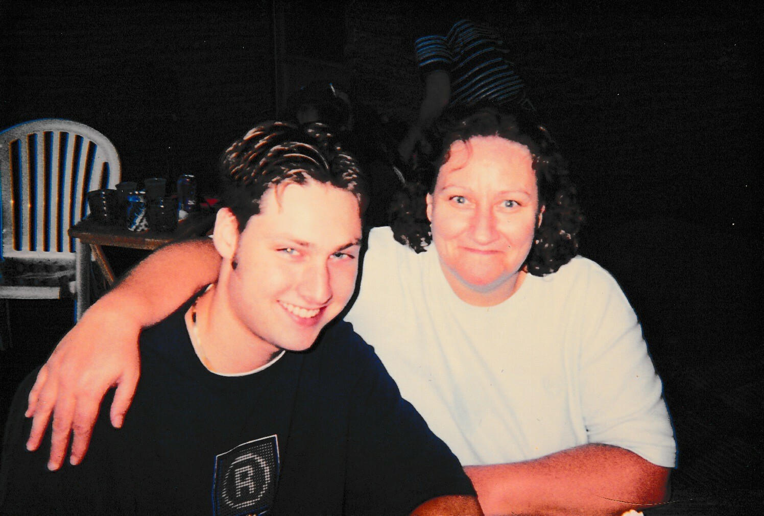 Two friends smiling at the camera with arms around each other at a casual indoor gathering in front of a table with cups and chairs in the background.
