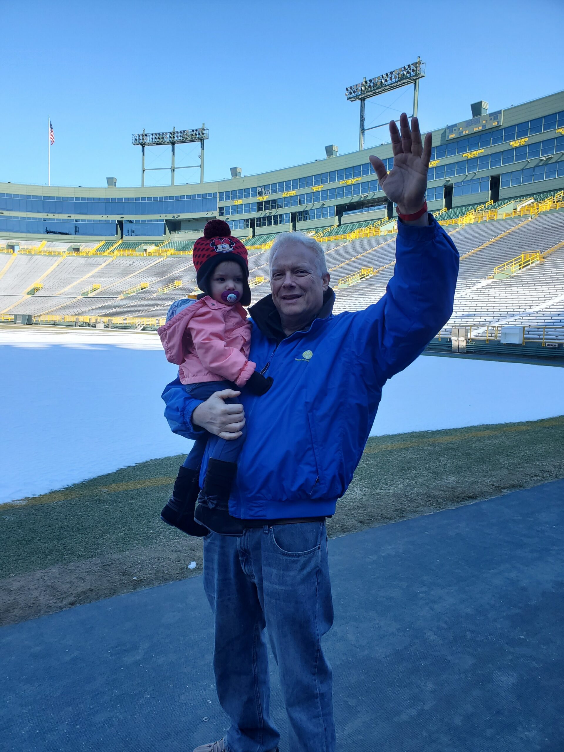 Man in a blue jacket holding a young child in a pink coat, while raising his hand and posing on a large empty stadium field under a clear blue sky.
