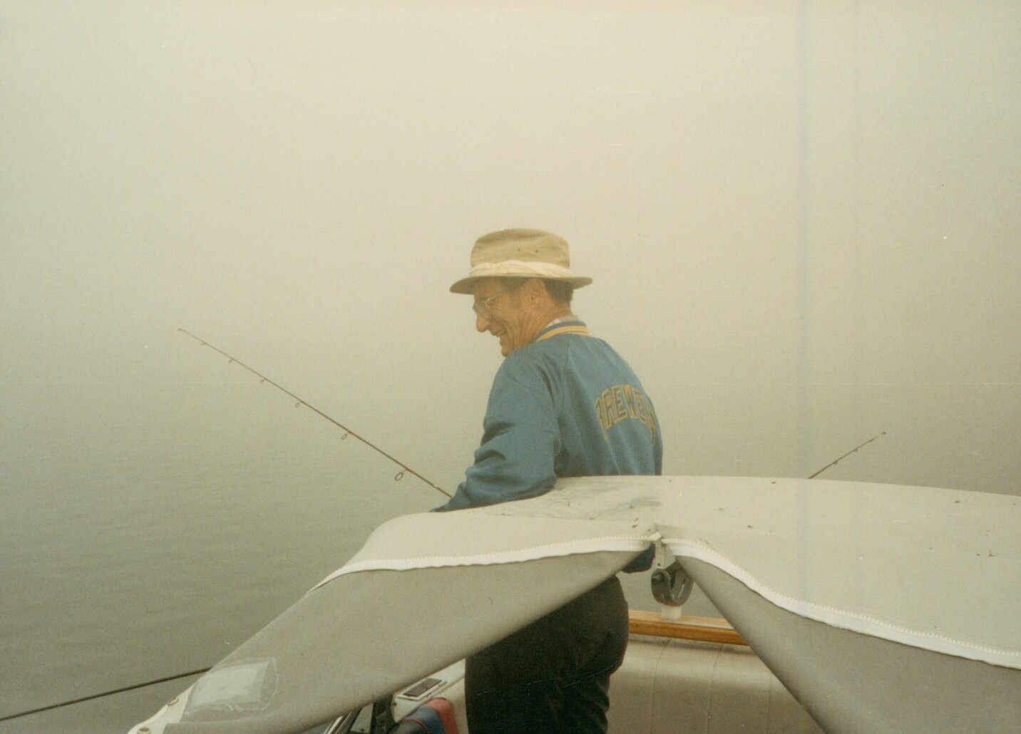 Man in a tan hat and blue jacket standing on a boat, fishing rod extended into calm water as he looks to the side.
