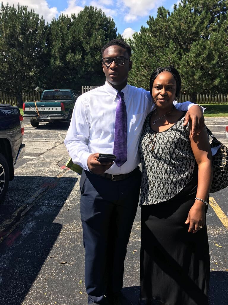 Young man in a white dress shirt and purple tie with his arm around a woman in a patterned top, posing in a sunny parking lot with trees in the background.