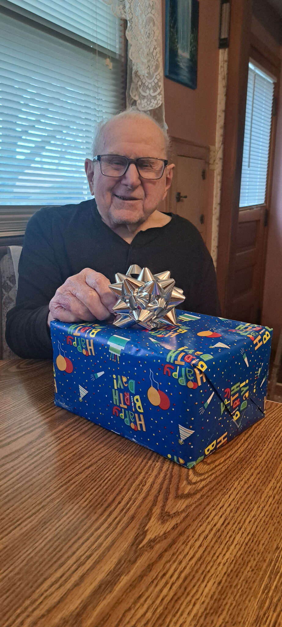 Older man with glasses smiling at a blue birthday gift with a large silver bow on a wooden table indoors