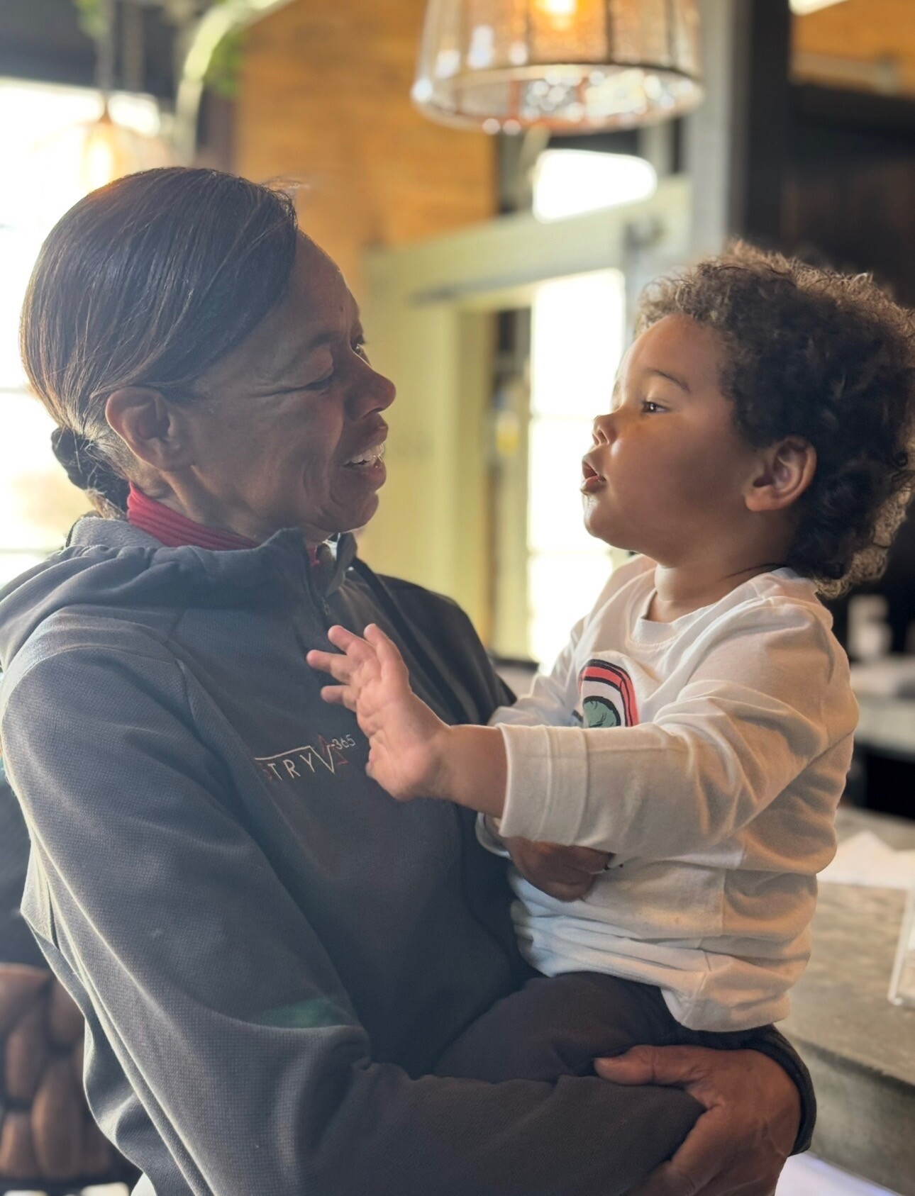 Adult woman holding a toddler, both facing each other and talking in a cozy indoor space.