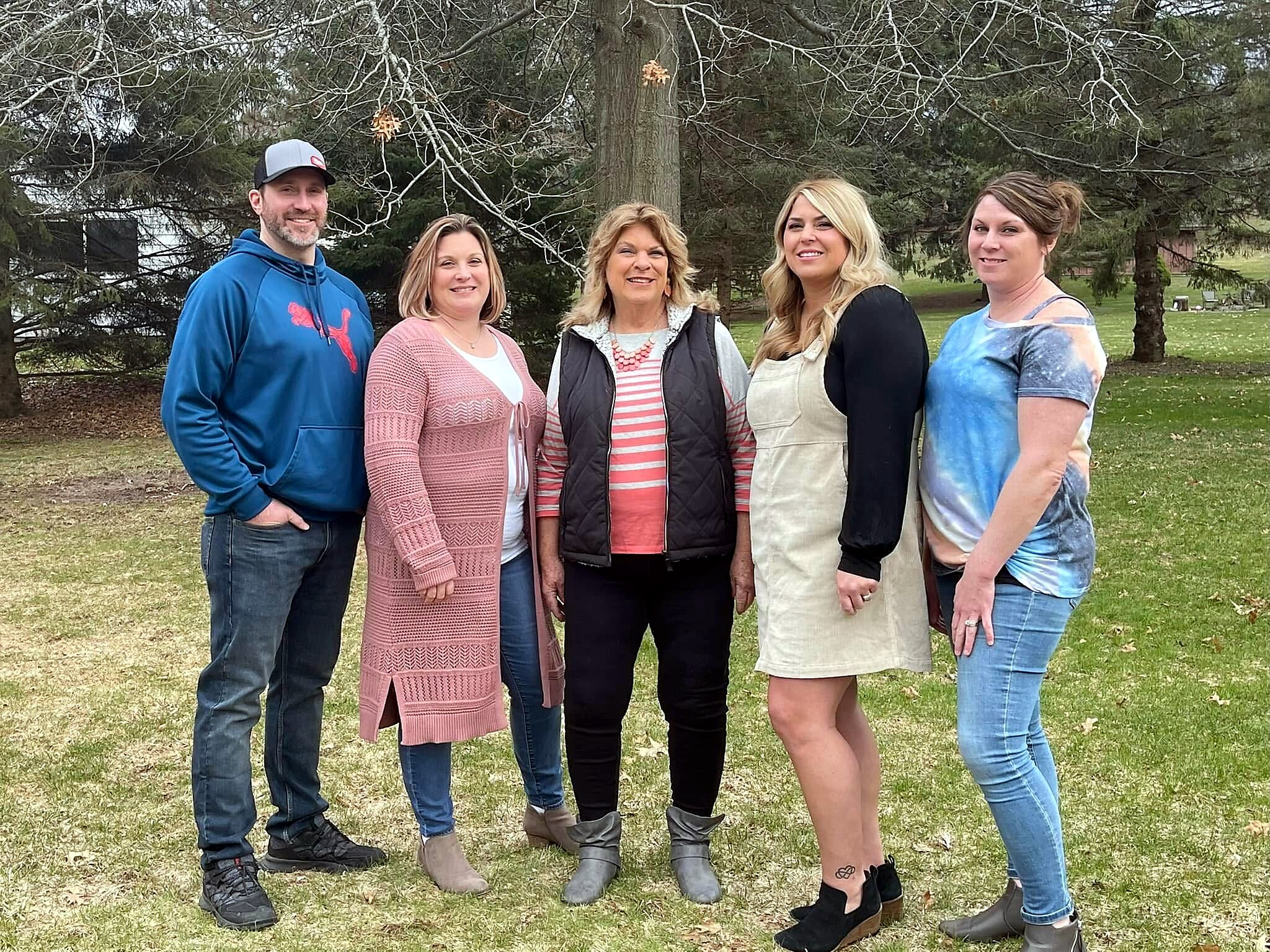 Five adults posing outdoors in a park, smiling, with trees in the background.