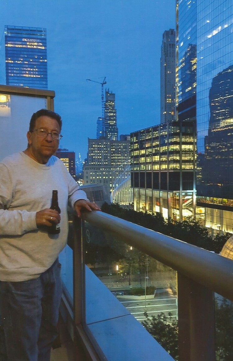 Man standing on a balcony with a beer bottle, city skyscrapers glowing at dusk behind a glass railing.