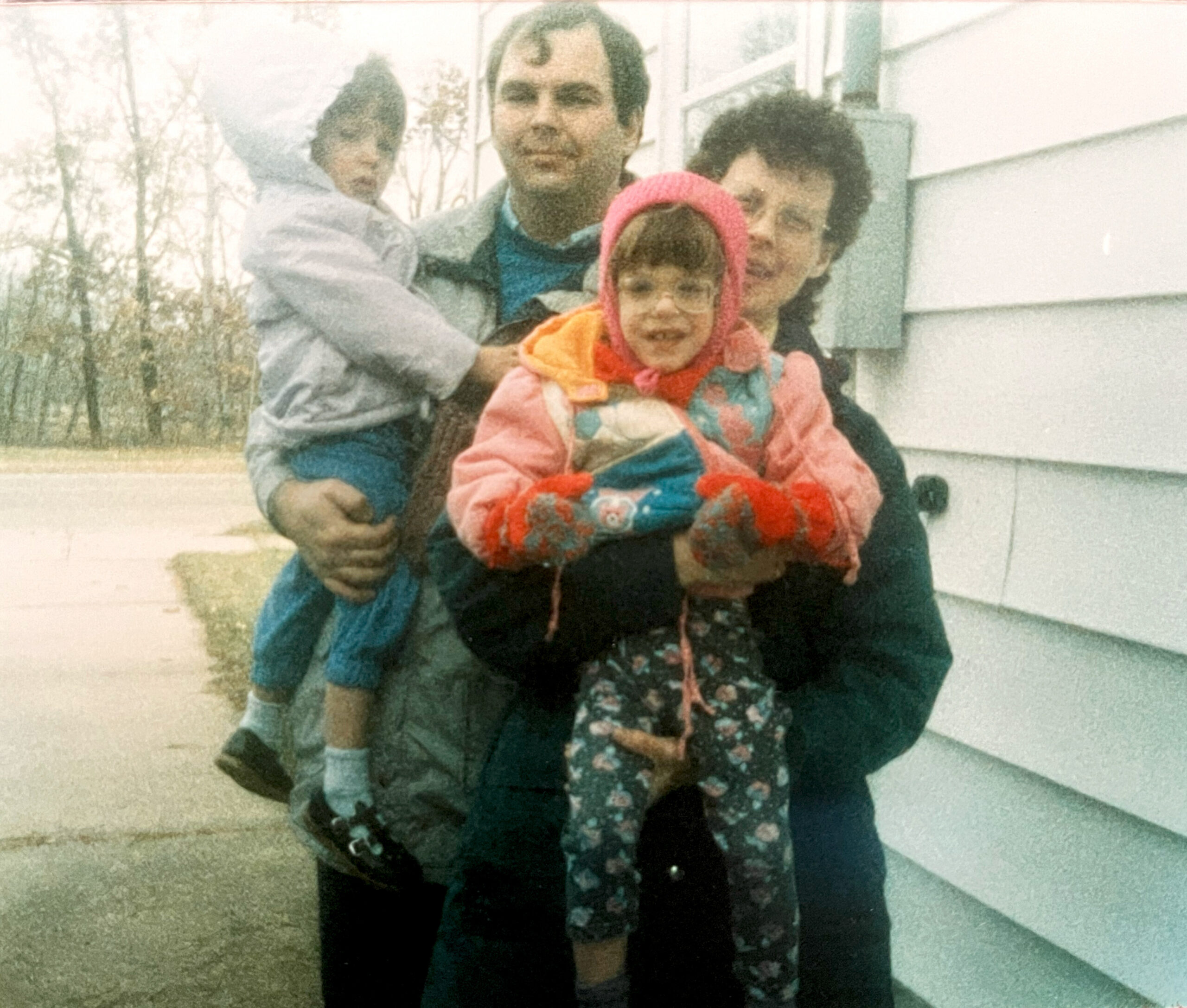 A small family posing outside a light-colored house; a child in a pink hood sits in a parent's arms, all smiling at the camera.