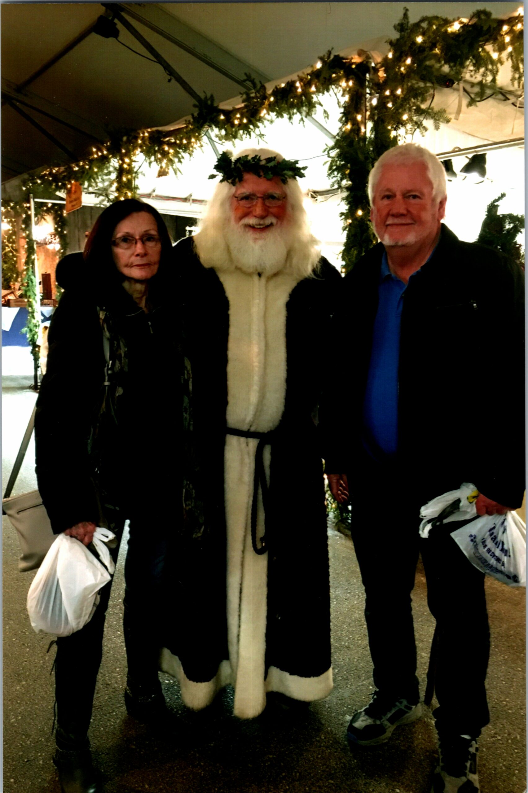 Three people pose together under festive lights; center figure wears a long white Santa-like robe and a leafy crown of greenery.