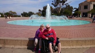 Two elderly people sit together on the edge of a circular fountain in a sunny park.